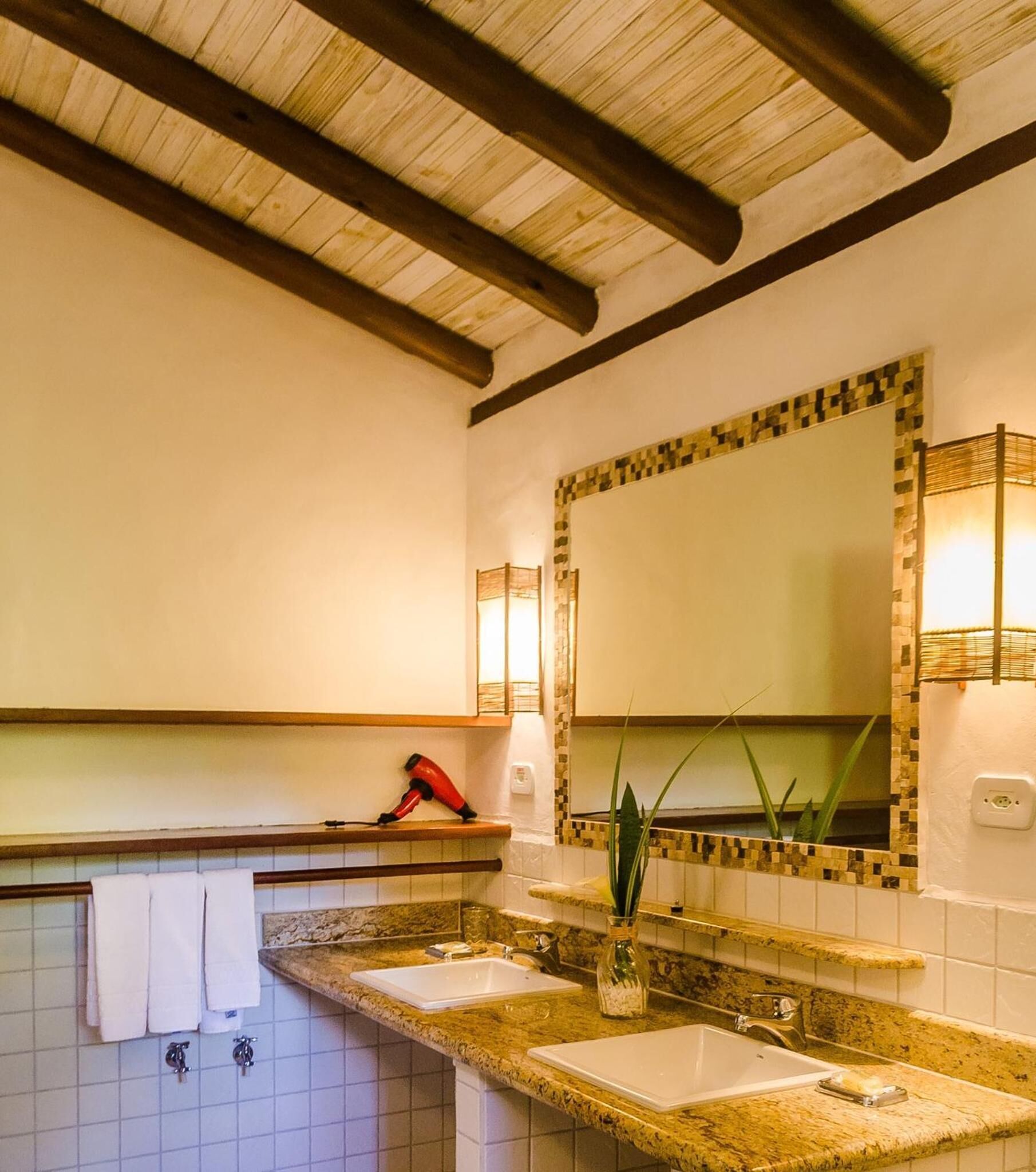 Un baño con techo de madera, dos lavamanos con encimeras de granito, un espejo con marco de mosaico y un secador de pelo rojo sobre el mostrador.