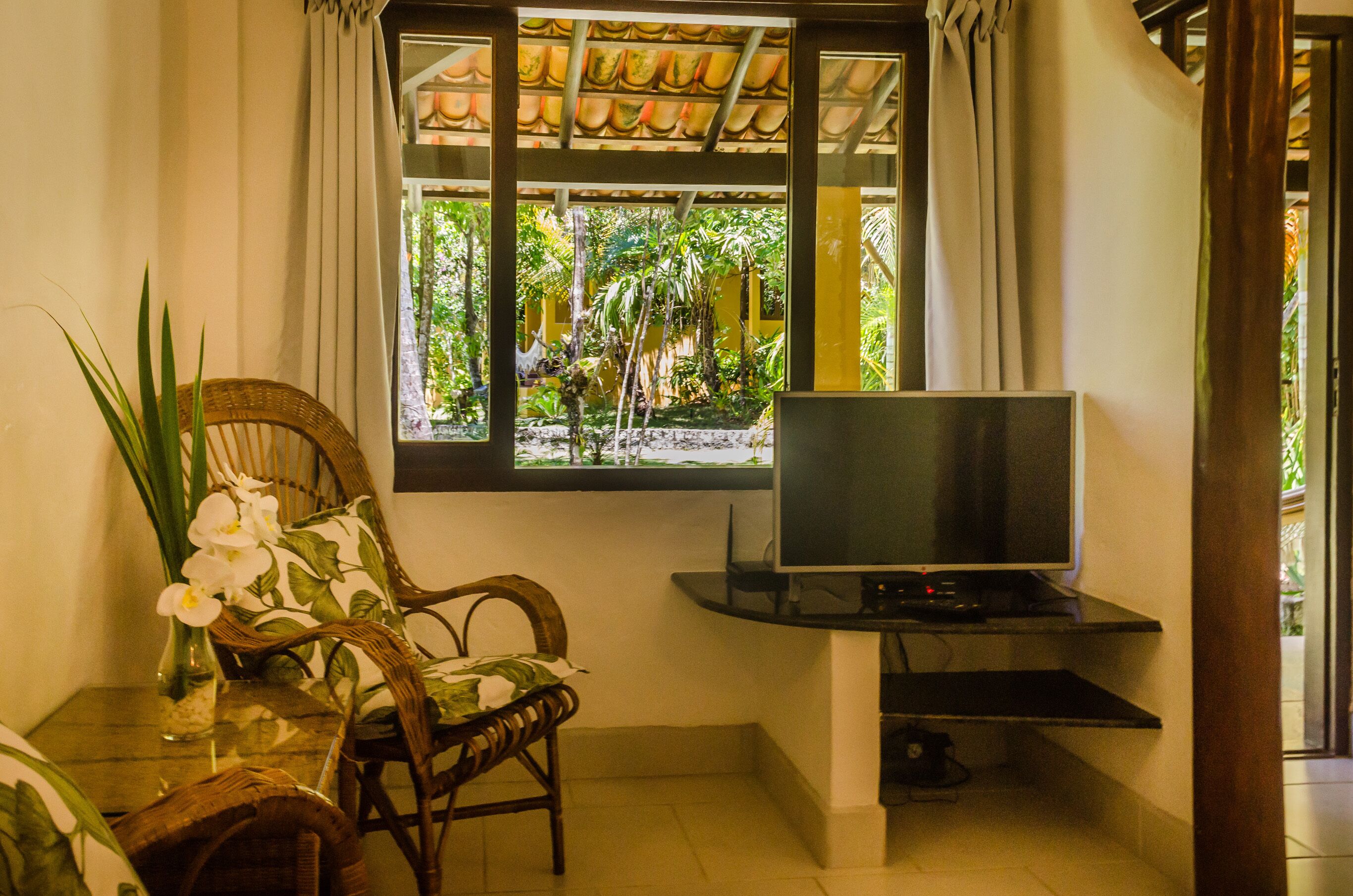 A room with a TV on a stand, a wicker chair, a small table with a vase of flowers, and a window with a view of greenery.