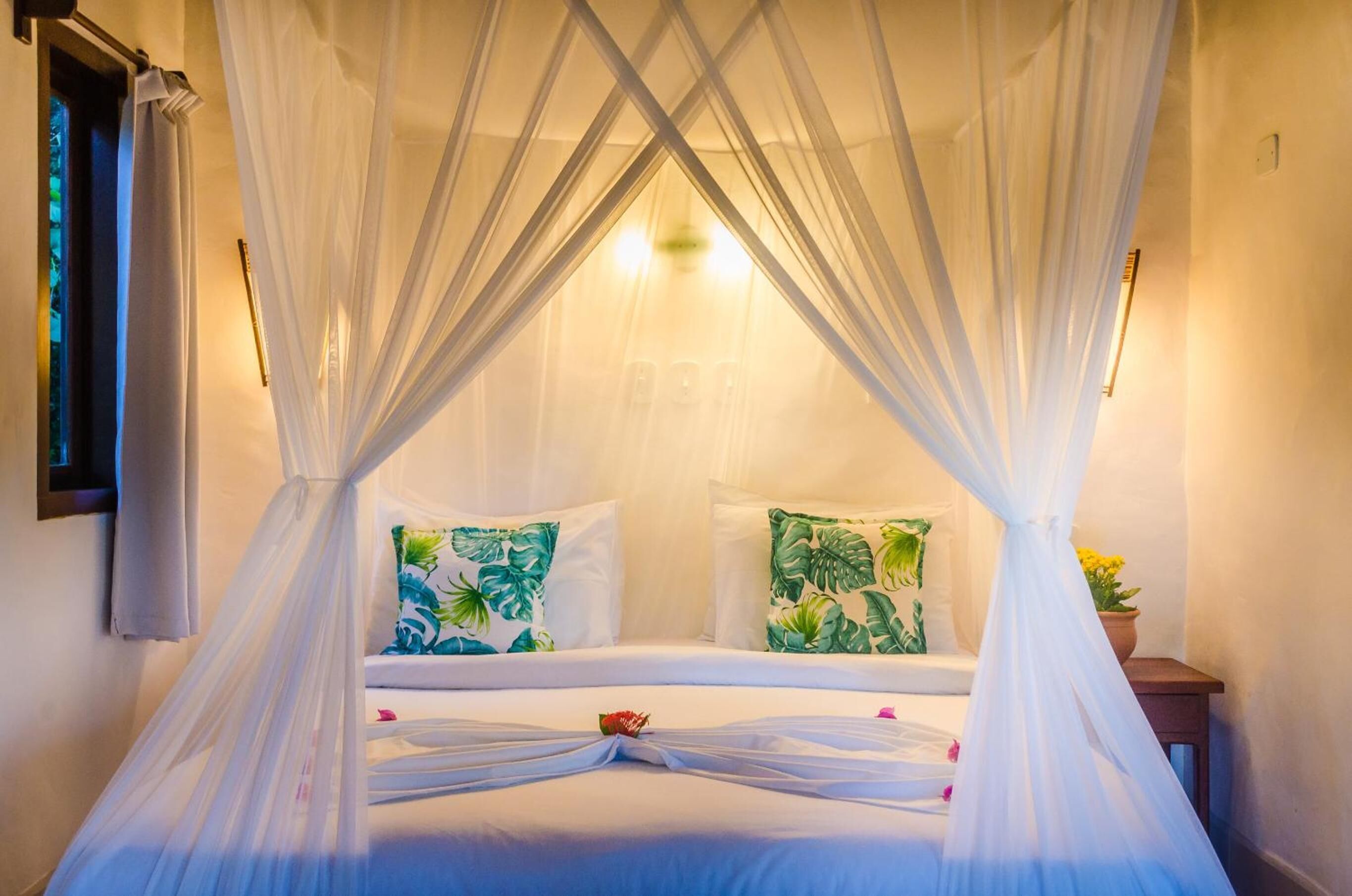 A bed with a canopy, two pillows with tropical leaf patterns, and a bedside table with a potted plant.
