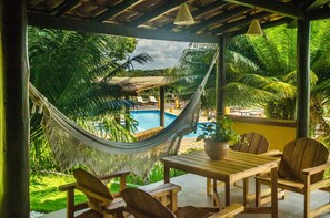 Una hamaca, una mesa de madera y sillas con vistas a una piscina, palmeras y una estructura de techo de paja.