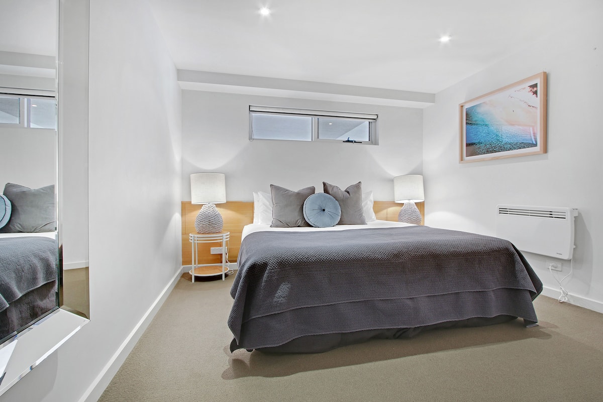 A modern bedroom with a large bed, a nightstand, a window, and a painting on the wall.