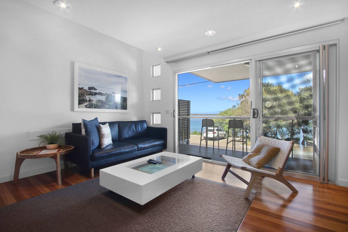 A modern living room with a dark blue sofa, a white coffee table, and a glass door leading to an outdoor area with a view of the sea.
