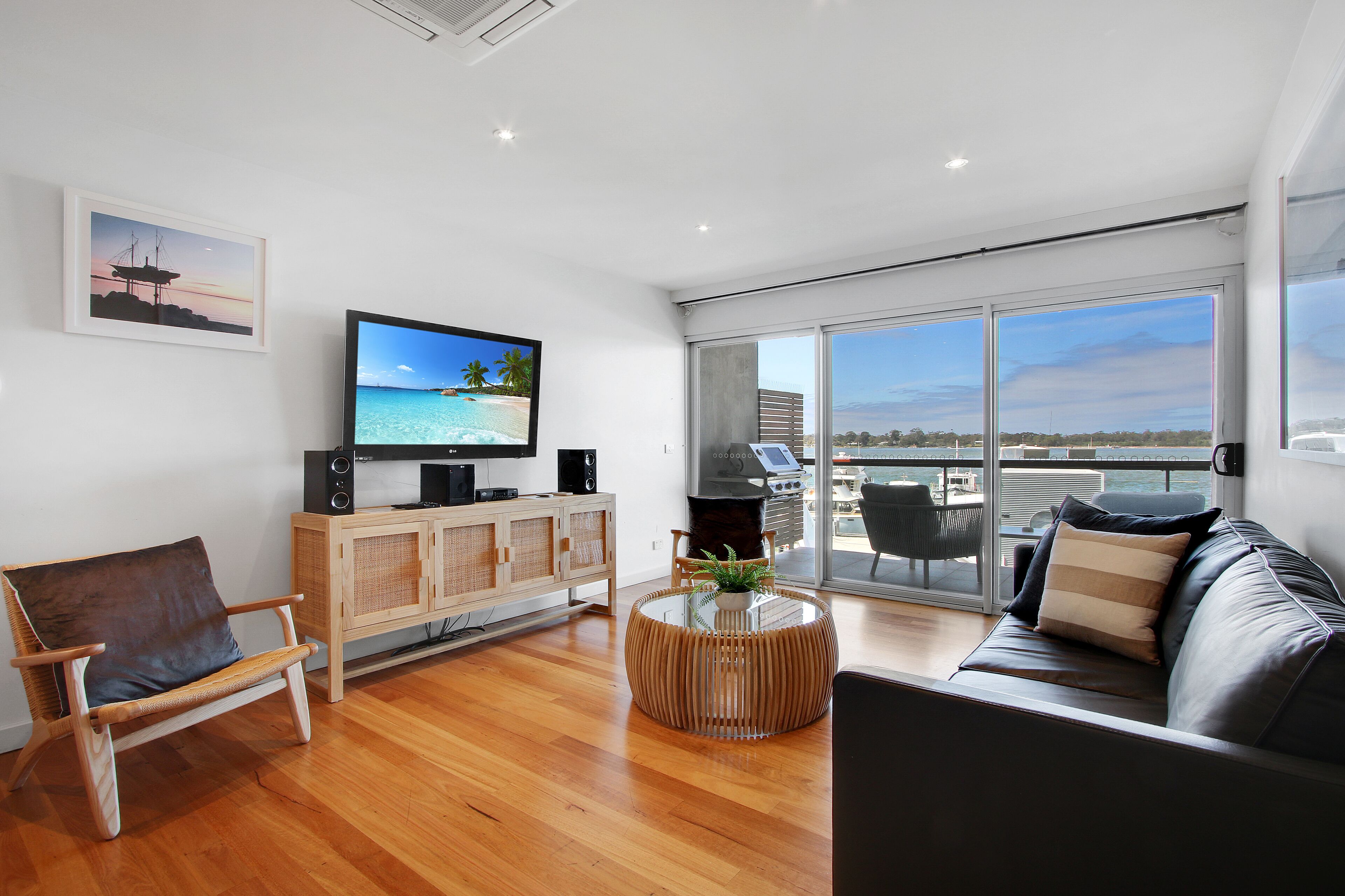 A modern living room with a wooden floor, a sofa, a TV, a wooden cabinet, and a large window with a view of the sea.