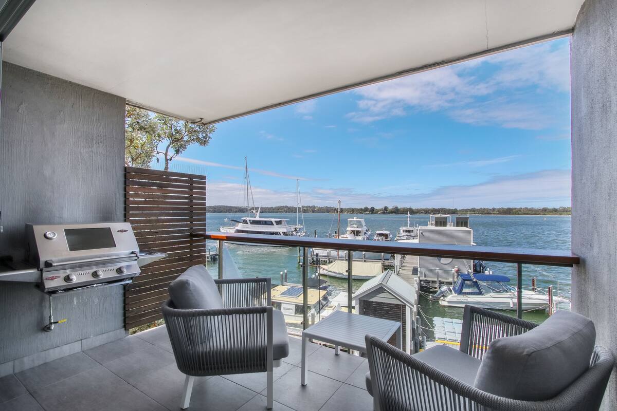 A balcony with a grill, two chairs, and a table overlooking a marina with boats.