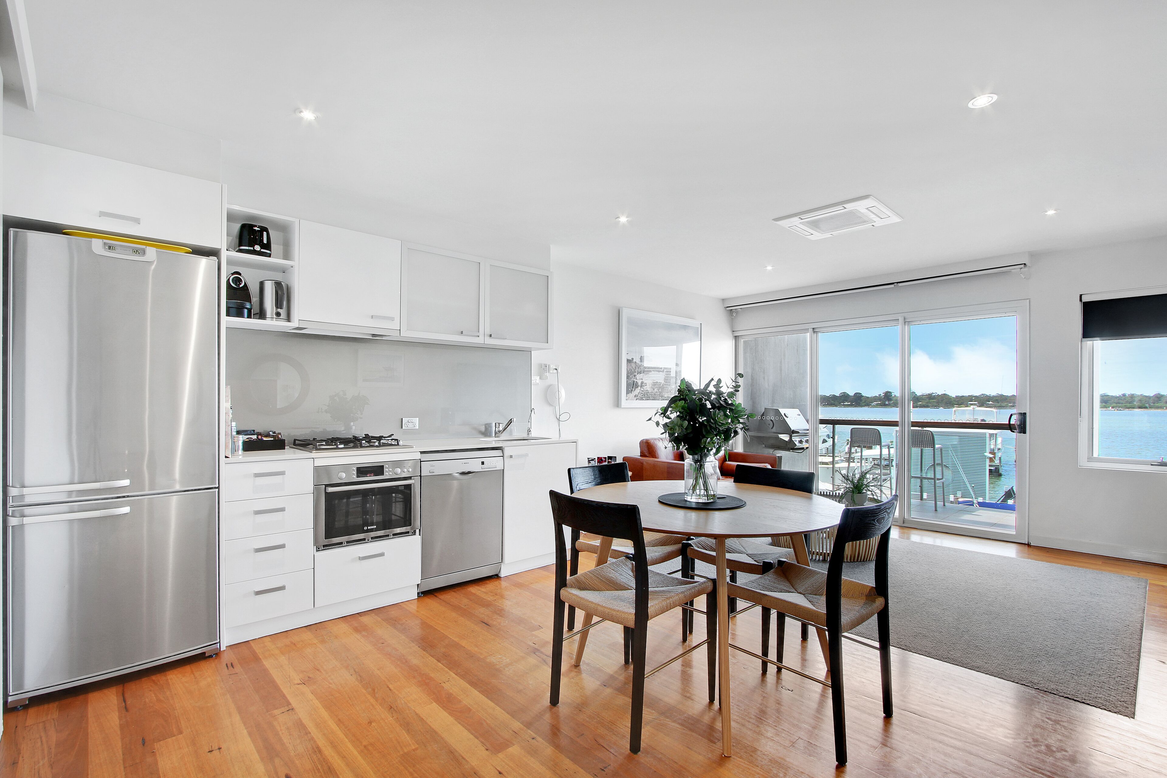 A modern kitchen with stainless steel appliances, a dining area with a round table and chairs, and a view of a body of water through large windows.