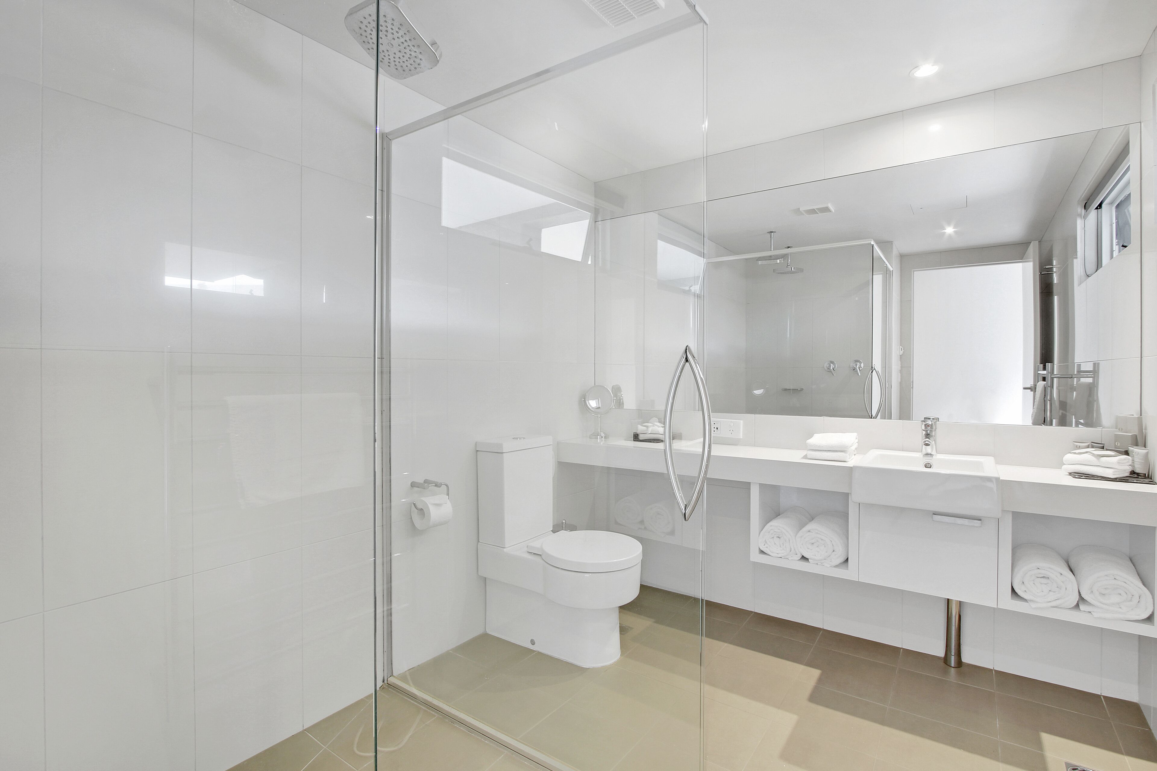 Une salle de bain moderne avec une douche à parois de verre, des toilettes blanches, un lavabo avec un miroir et des étagères avec des serviettes.