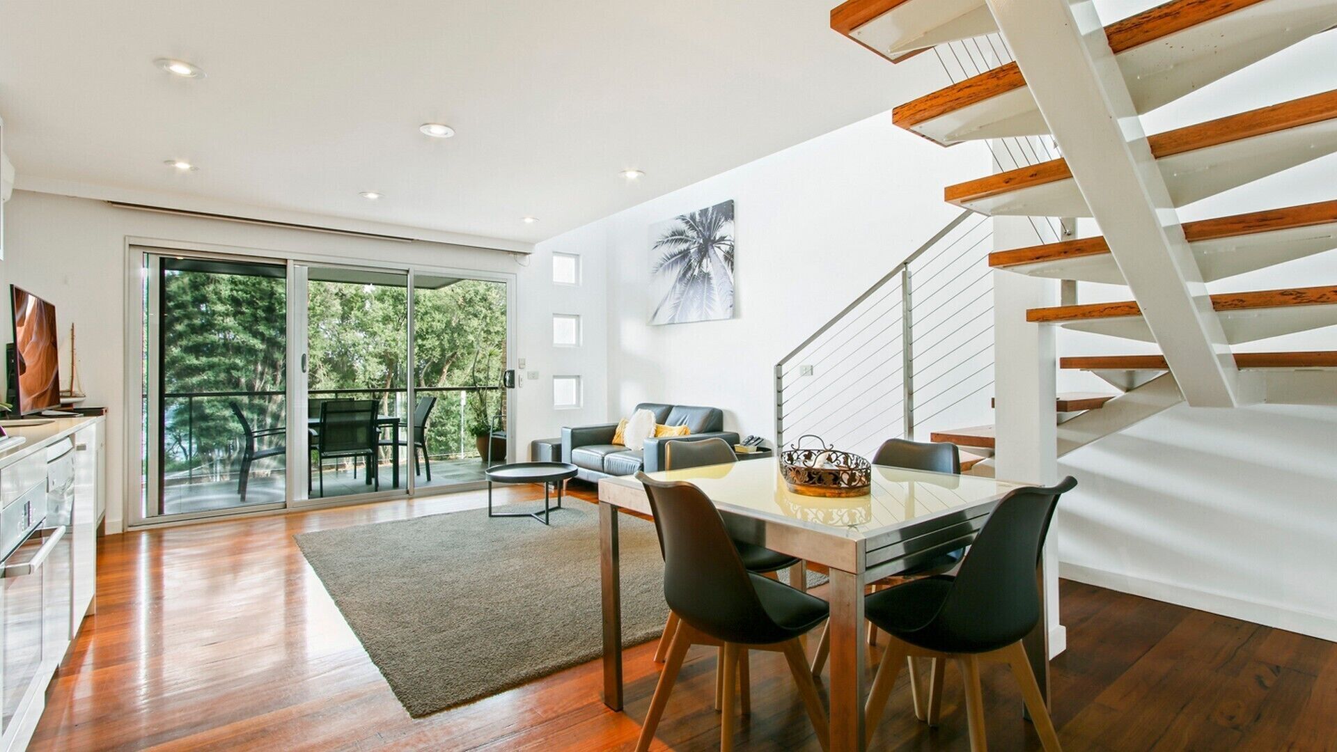 Un interior moderno con comedor, una escalera y un gran ventanal con vistas a árboles.