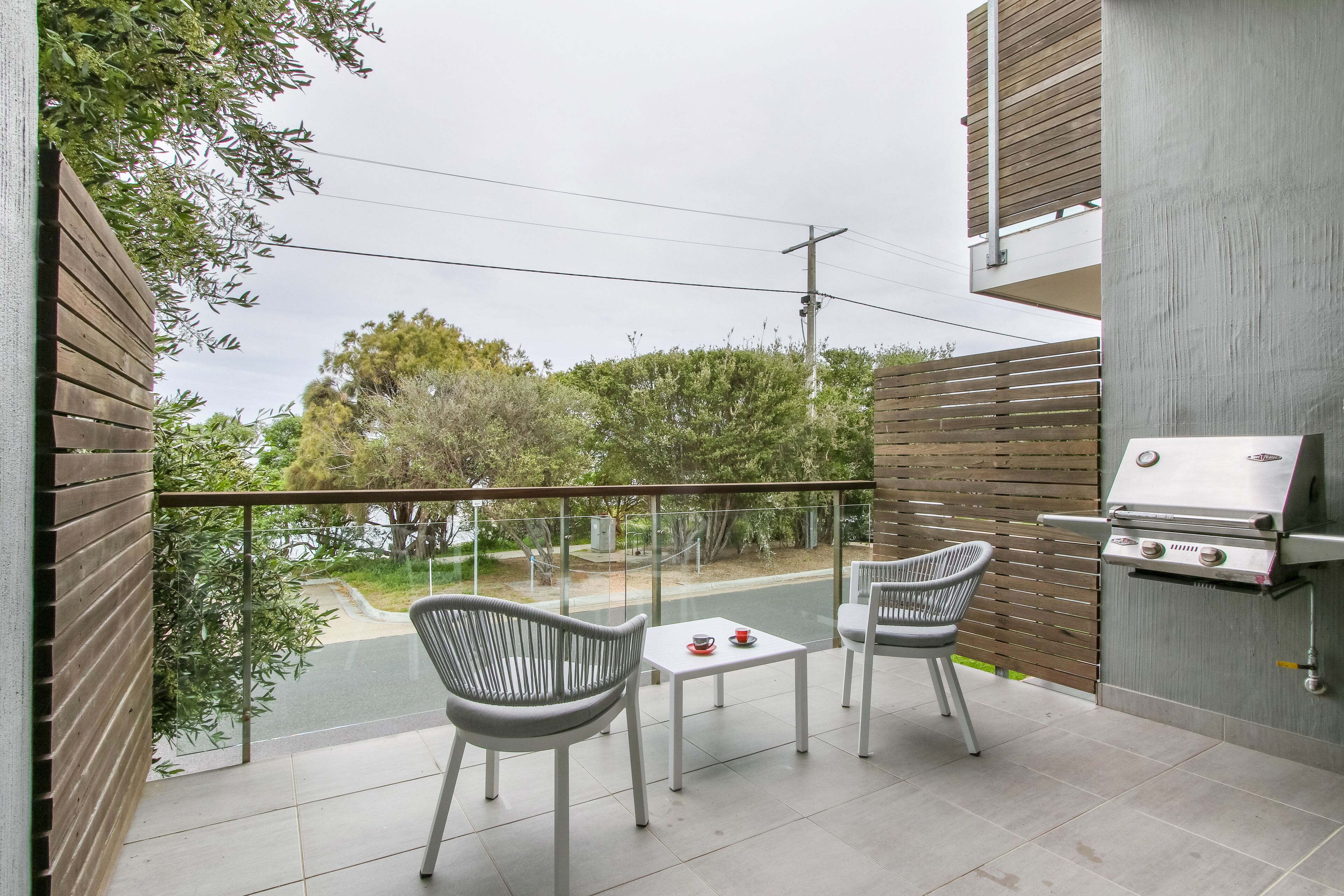 A balcony with two chairs and a table, a barbecue grill, and a view of the street and trees.