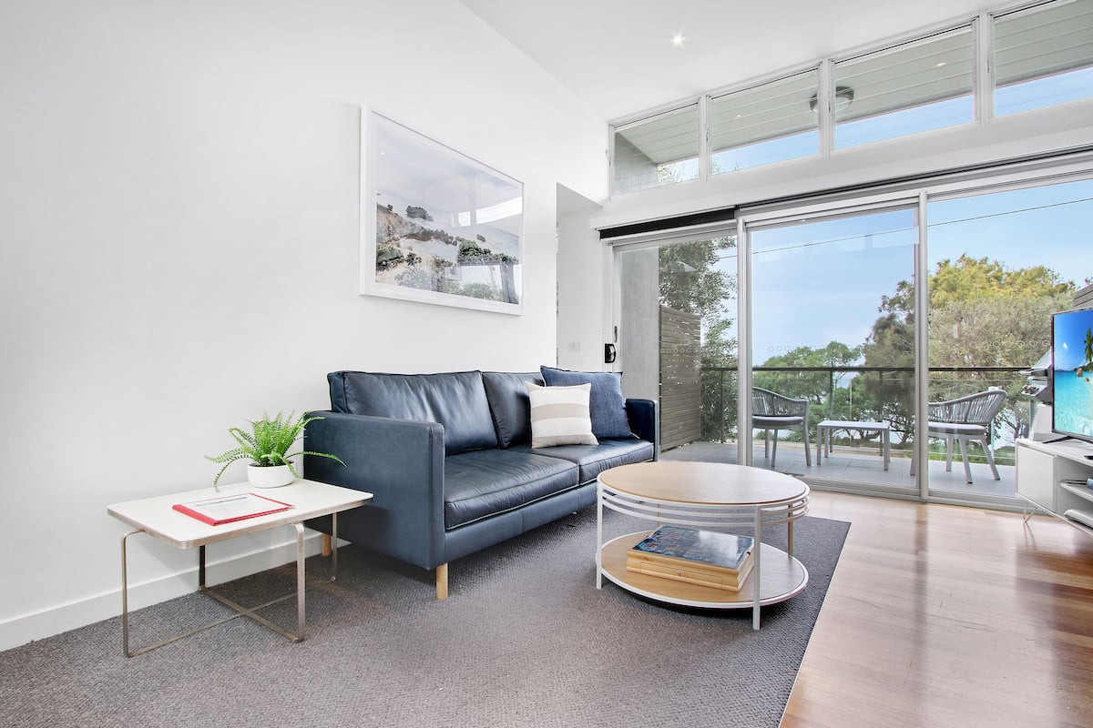 A modern living room with a leather sofa, a small side table, a round coffee table, and a large window offering a view of the outdoors.