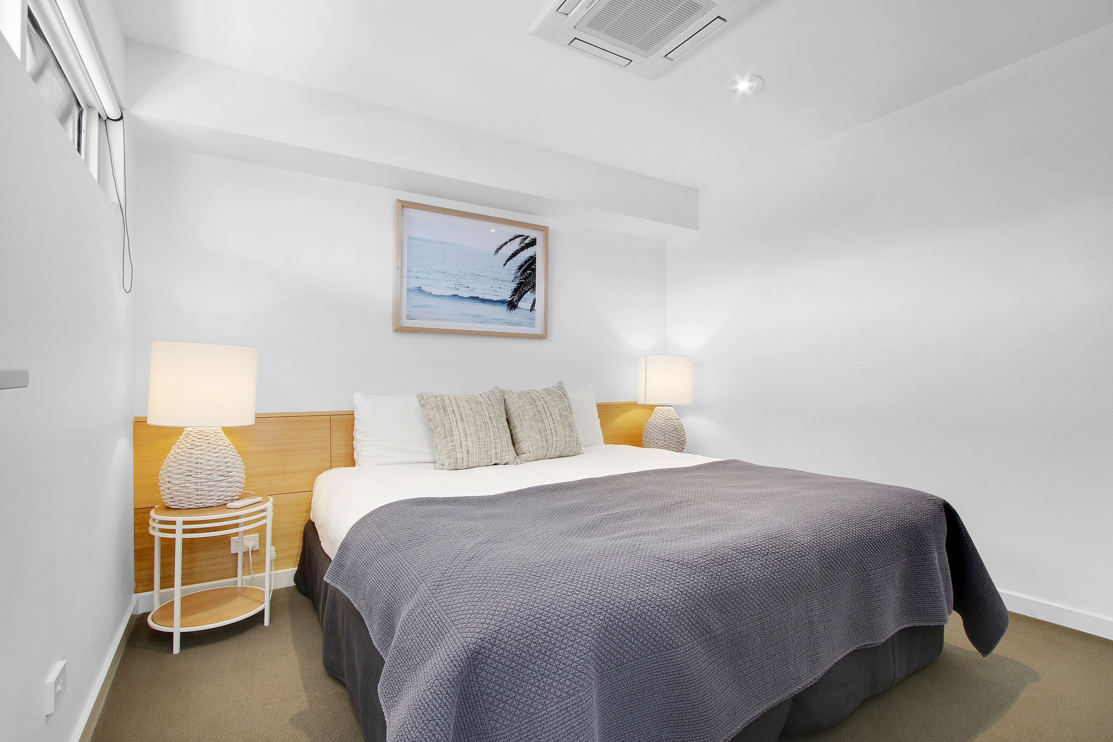 Habitación de hotel impecablemente ordenada, con una cama, dos lámparas de noche, un cuadro enmarcado con una escena de playa y un cabecero de madera.