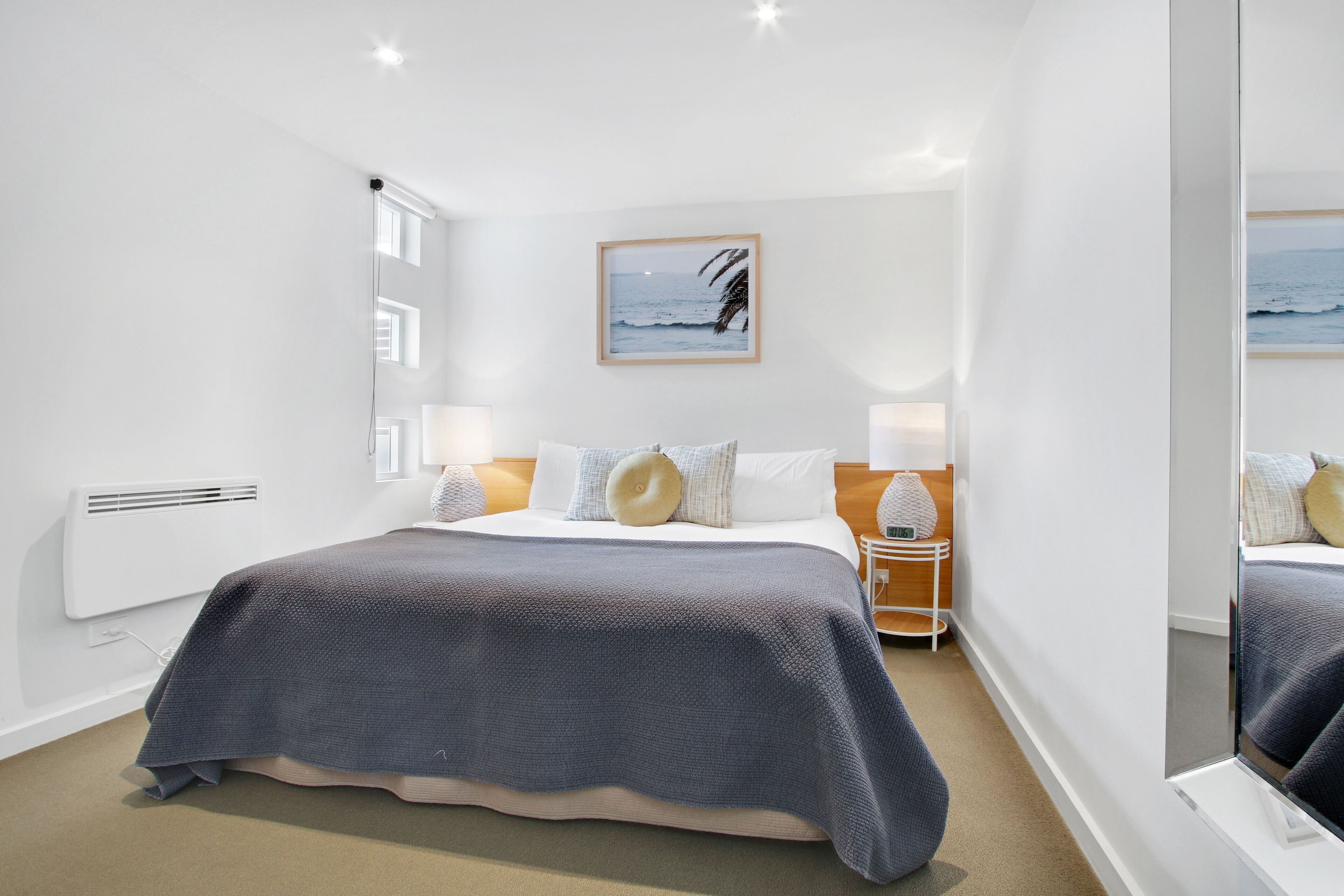 A modern hotel room with a large bed, a bedside table with a lamp, a wall-mounted air conditioner, and a framed picture of the ocean.