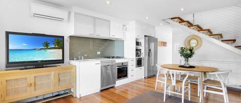 A modern kitchen with a large TV, wooden cabinets, and a dining area with white chairs.