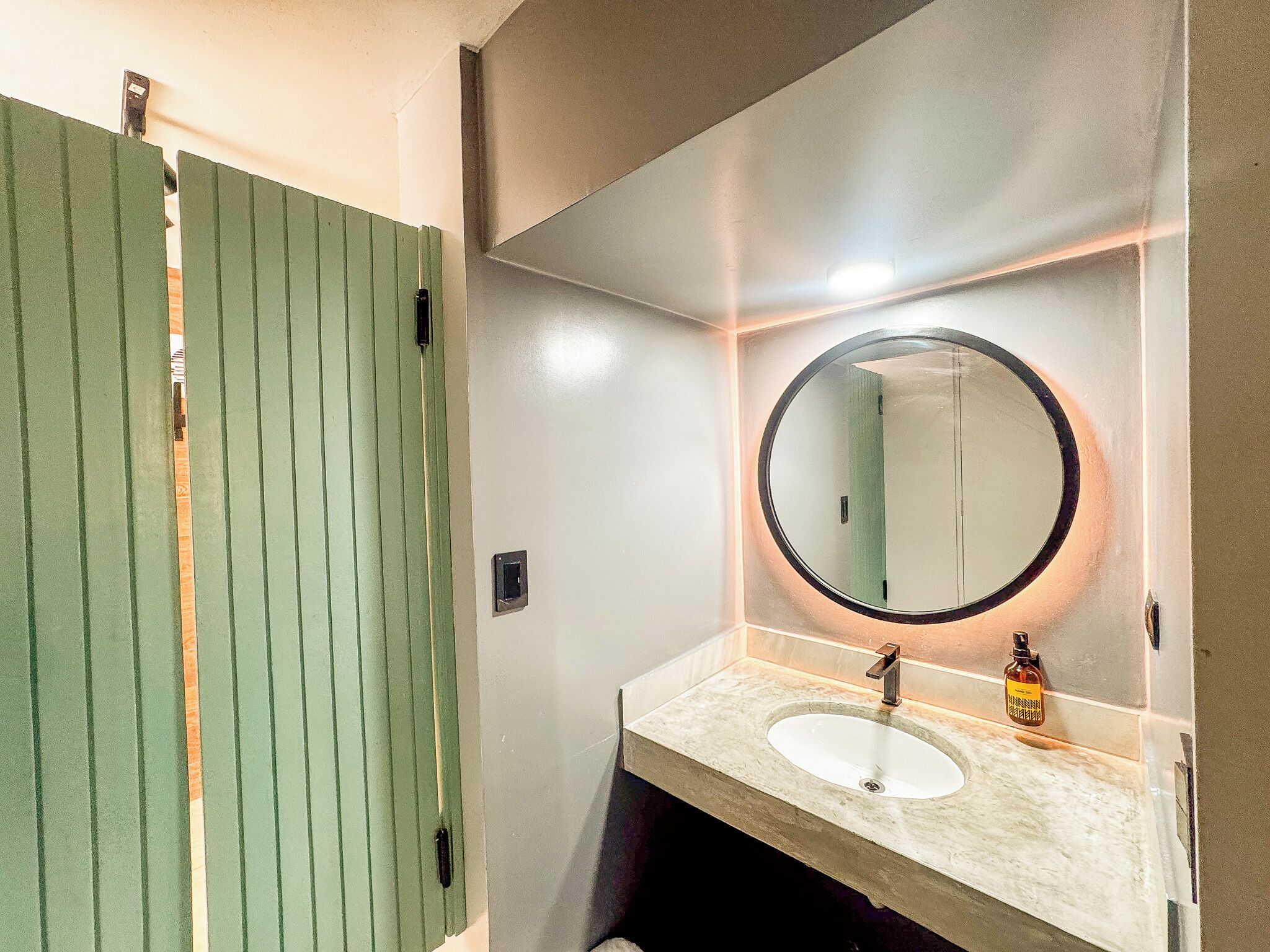 A bathroom with a green door, a round mirror, a sink, and a bottle of hand soap.