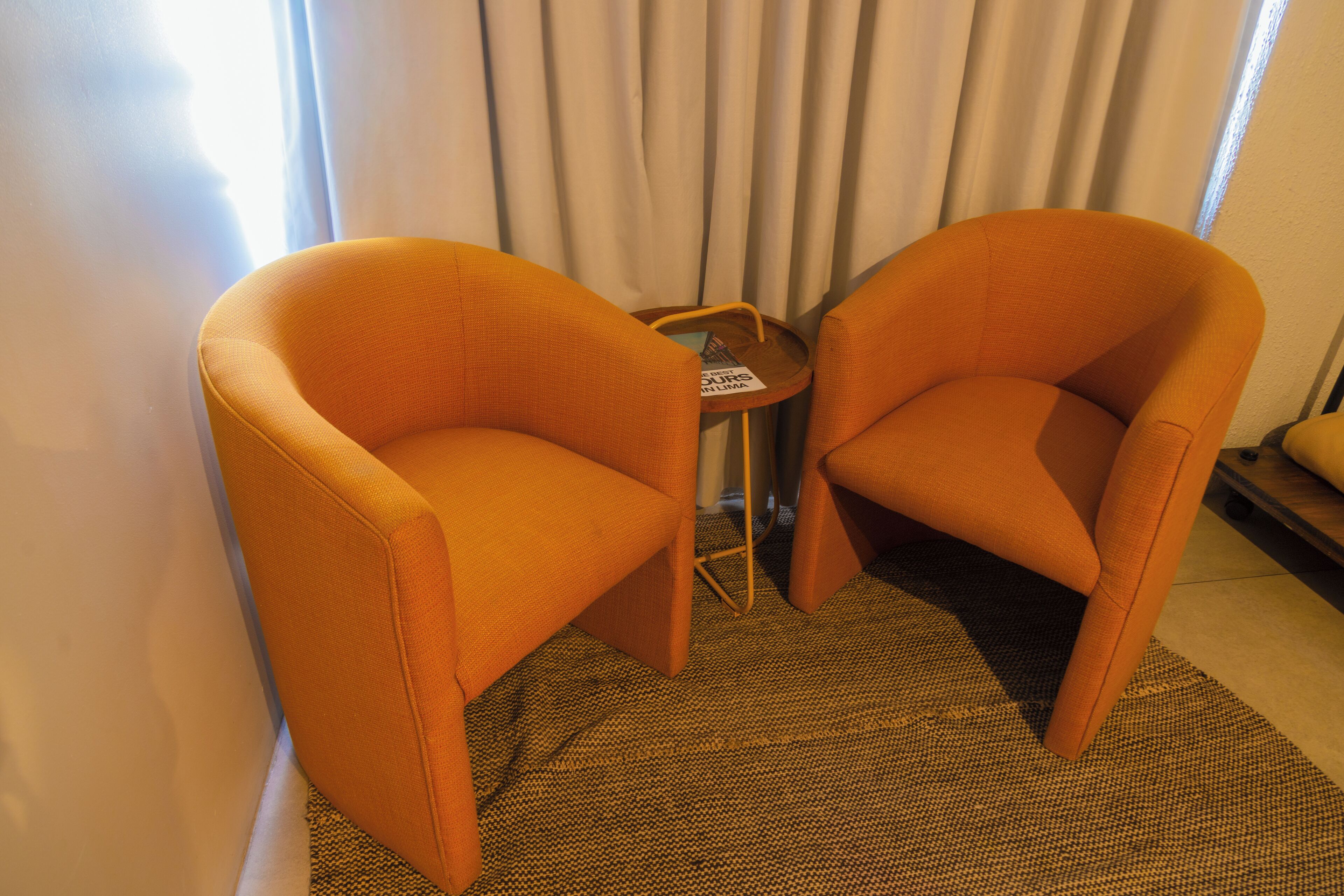 Dos sillones naranjas, una mesita con una revista y una alfombra estampada en una habitación con cortinas.