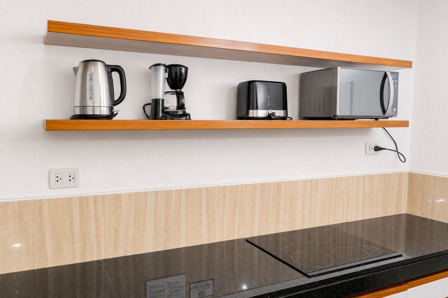 A kitchen with a microwave, coffee maker, and kettle on a wooden shelf, a black countertop, and a wall socket.