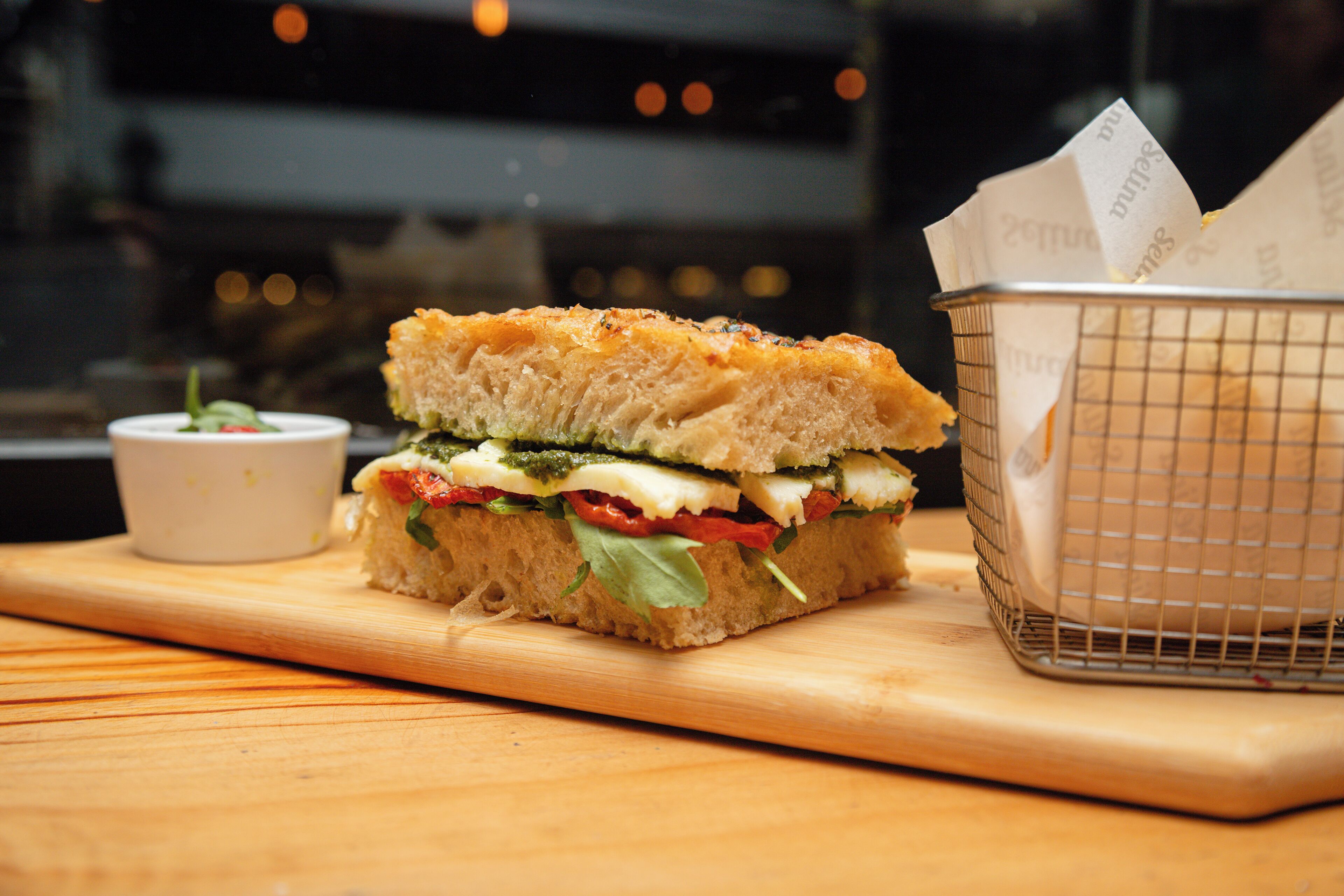 A sandwich on a wooden board with a small bowl of sauce and a basket with napkins.