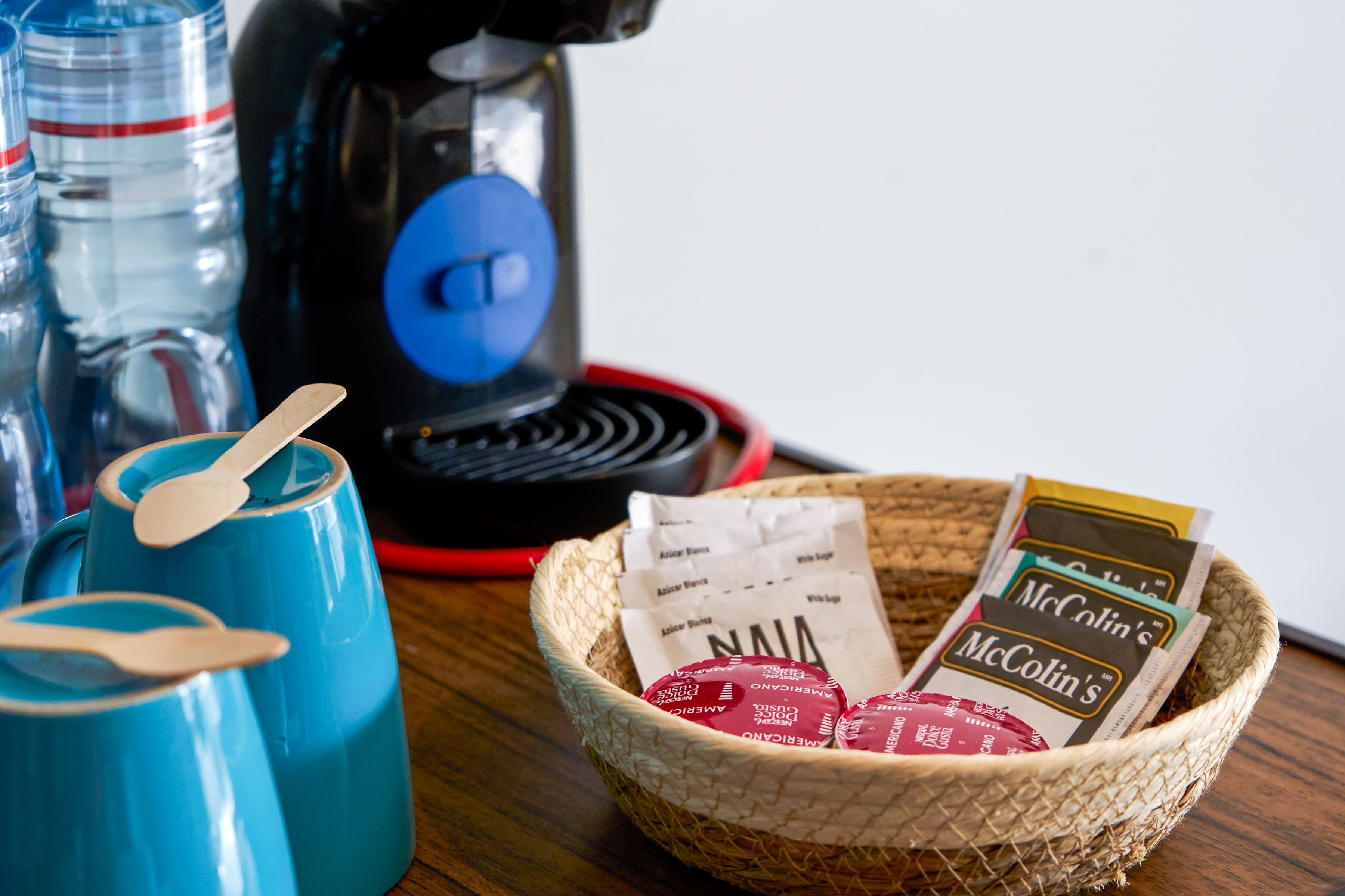 Une machine à café, des tasses bleues et un panier contenant des sachets de café McColin, le tout posé sur une surface en bois.