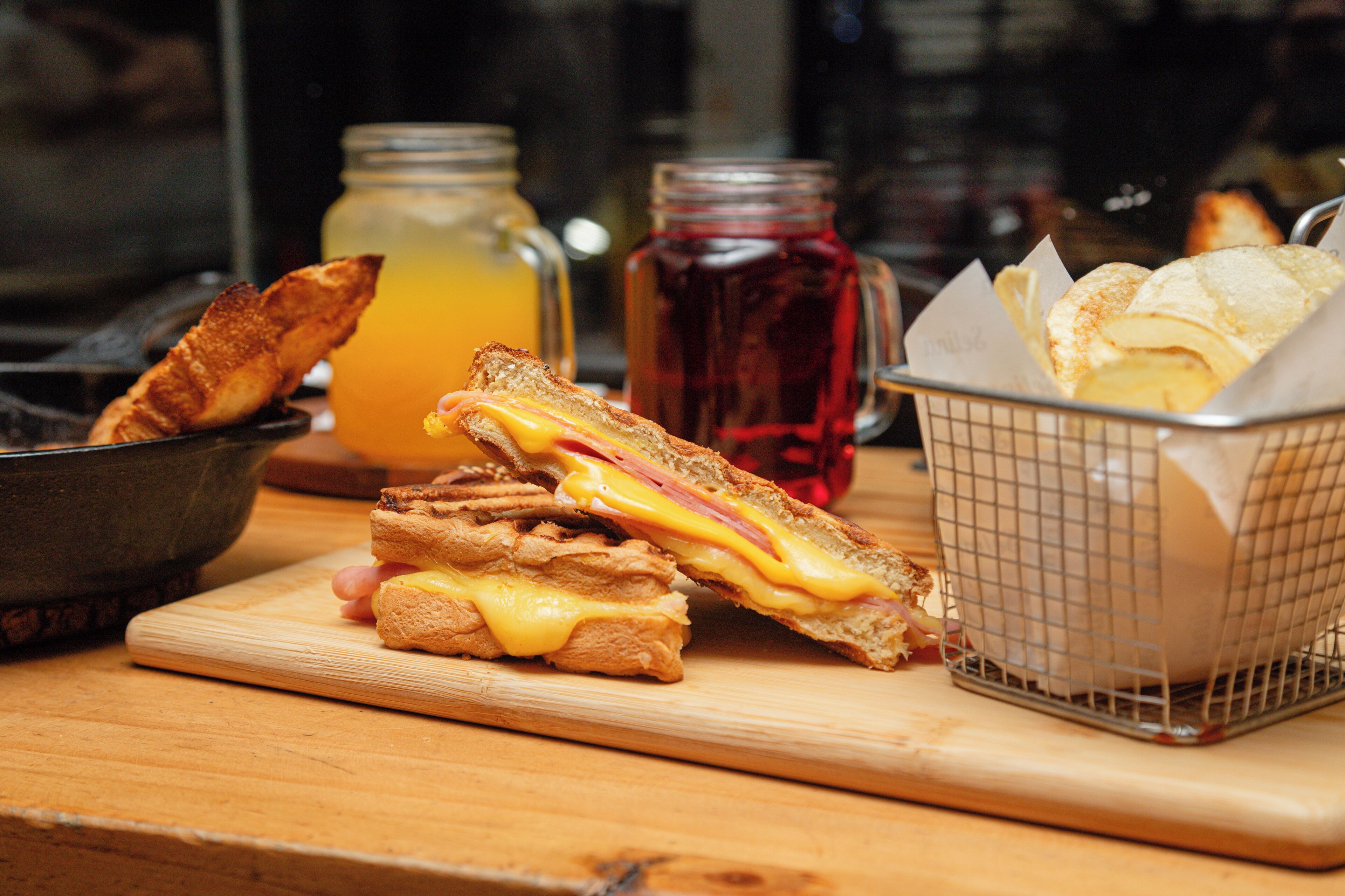 A sandwich cut in half on a wooden board, a jar of orange juice, a jar of red jam, and a basket of bread rolls.