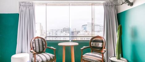 Una habitación con dos sillas de madera, una mesita redonda pequeña, un otoman blanco, una planta en maceta y un ventanal con cortinas.