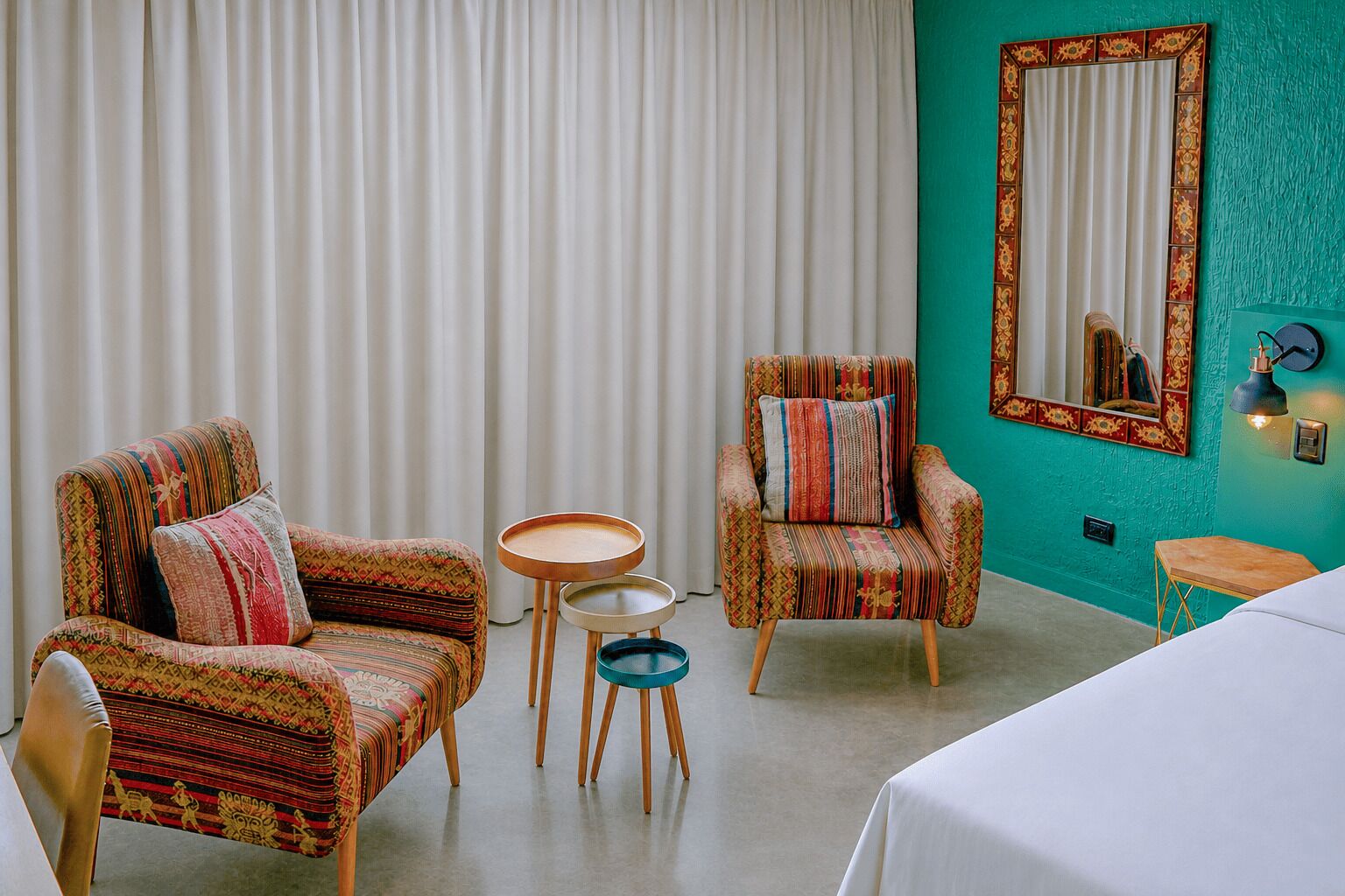 A room with two patterned armchairs, a small round table, a mirror with a decorative frame, and a bed.
