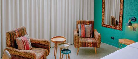 A room with two patterned armchairs, a small round table, a mirror with a decorative frame, and a bed.