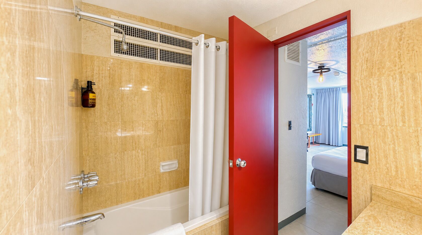 A modern bathroom with a glass-enclosed shower, a white bathtub, and a red door leading to a bedroom with a bed and a window.