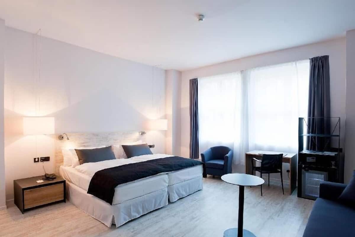 A hotel room with a bed, bedside tables, a desk with a chair, a TV, and large windows with curtains.