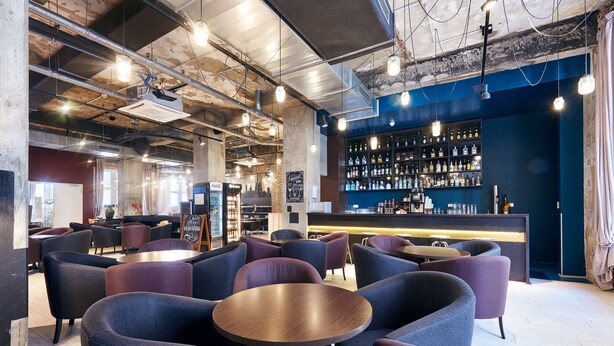 Un espace bar moderne avec des tables et des chaises rondes, un comptoir avec des étagères remplies de bouteilles, et des conduits apparents au plafond.
