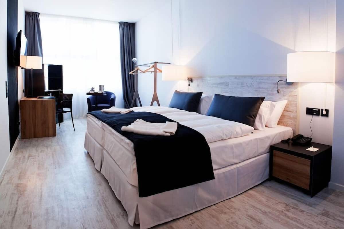 A hotel room with a bed, bedside table, lamp, TV, desk, chair, and a view of the outdoors.