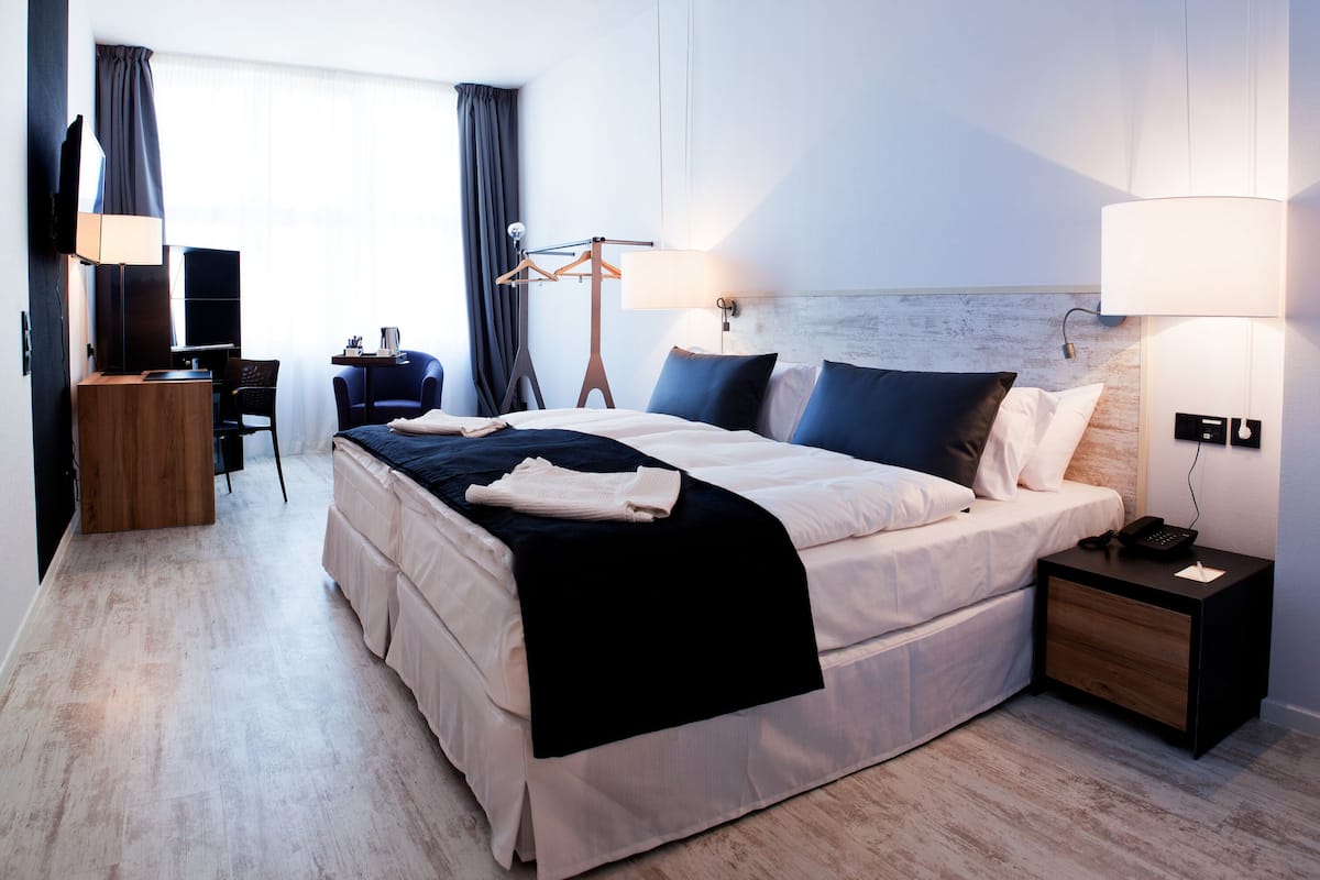 A hotel room with a bed, bedside table, lamp, TV, desk, chair, and a view of the outdoors.