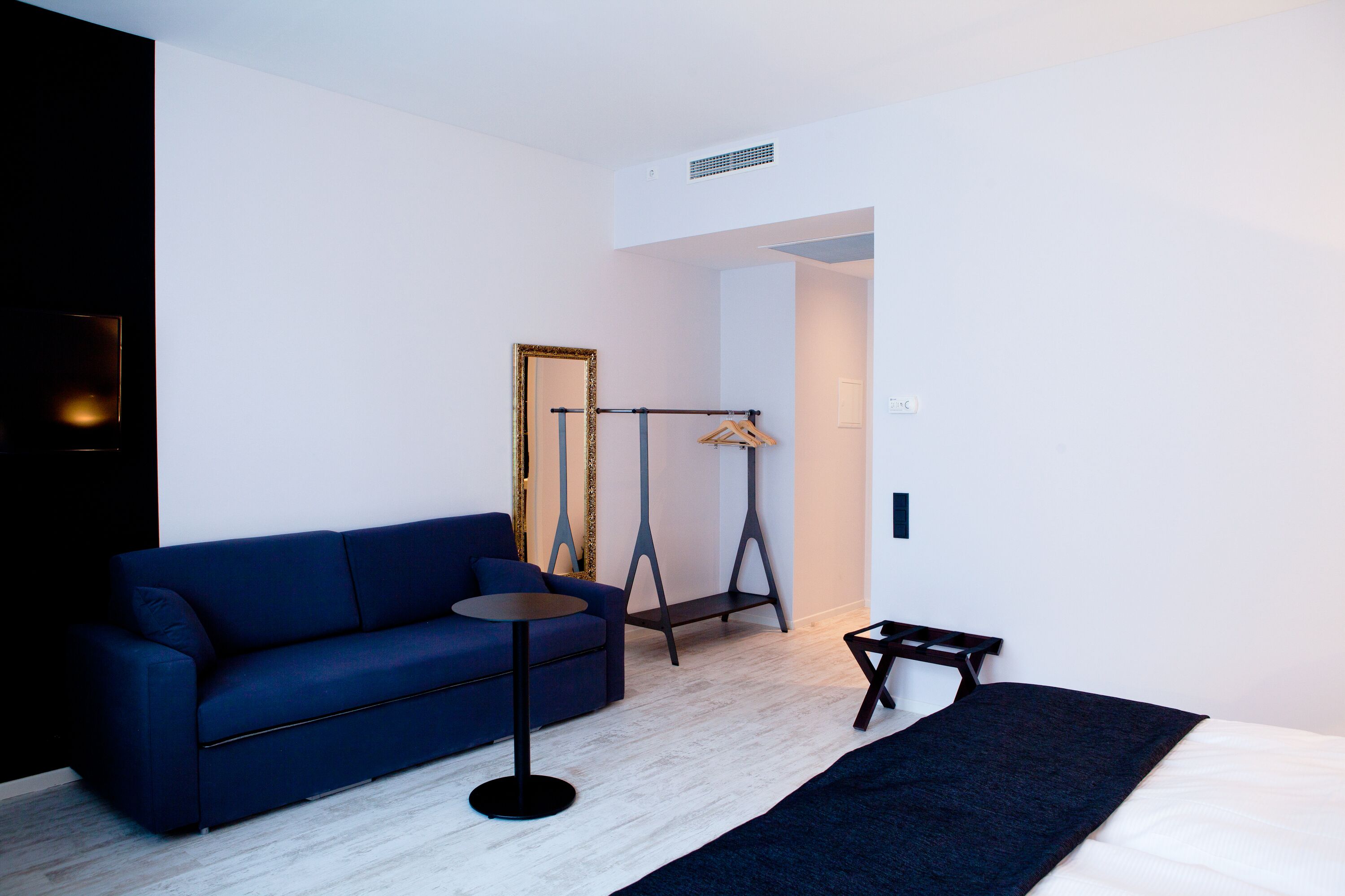 Una habitación de hotel moderna con un sofá azul, una pequeña mesa redonda, un perchero y una cama con un plaid azul oscuro.
