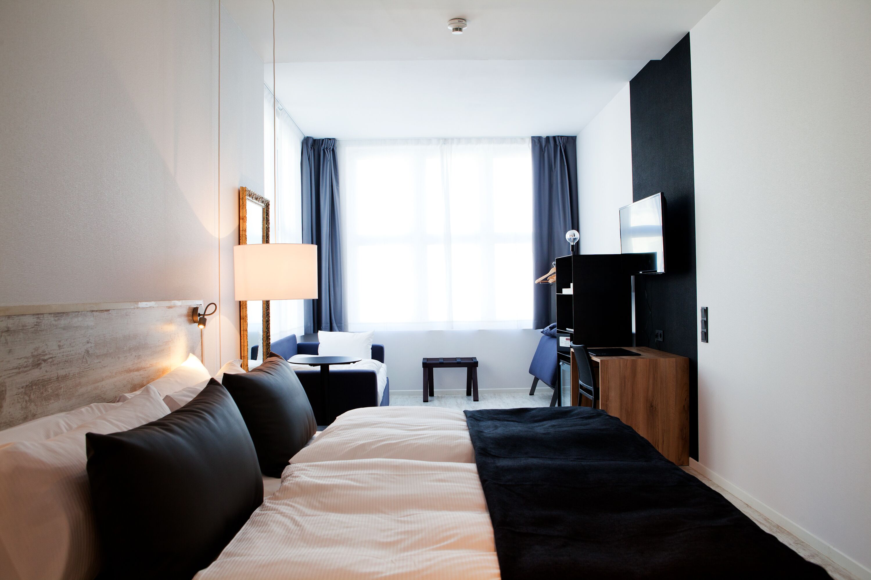 Ein Hotelzimmer mit Bett, Nachttisch, Lampe, Fernseher und kleinem Schreibtisch mit Stuhl.