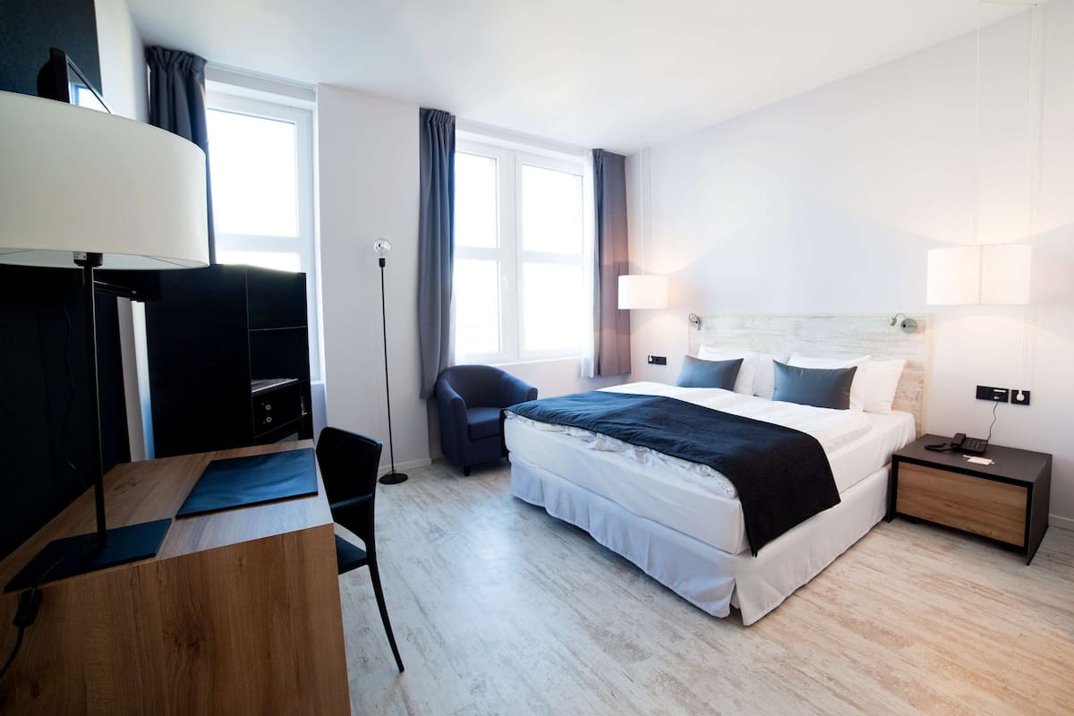 A modern hotel room with a bed, bedside table, lamp, and a desk with a chair.