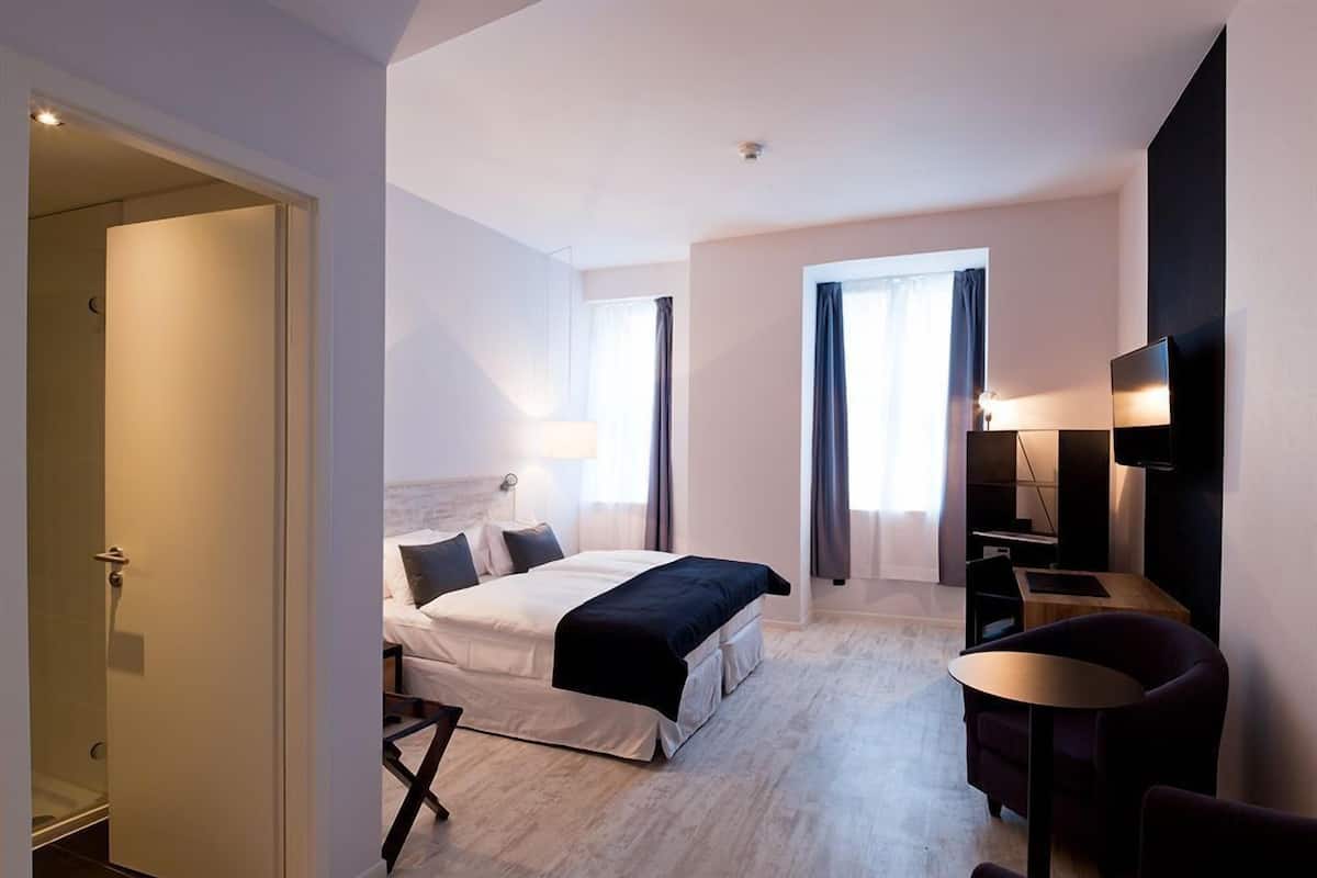 A hotel room with a bed, a desk, a chair, a TV, and a door leading to a bathroom.