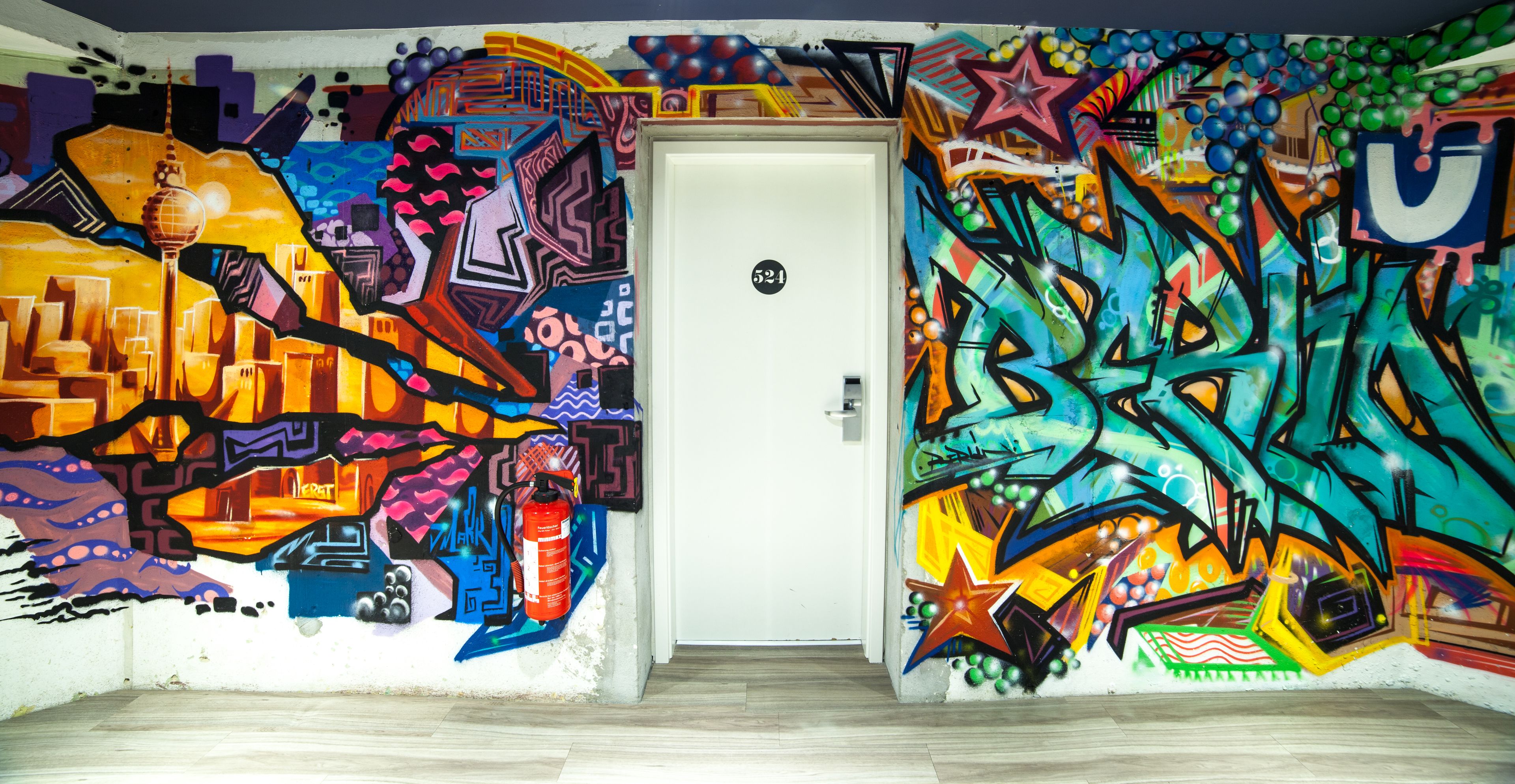 A room with vibrant graffiti art on the walls, a white door, and a wooden floor.