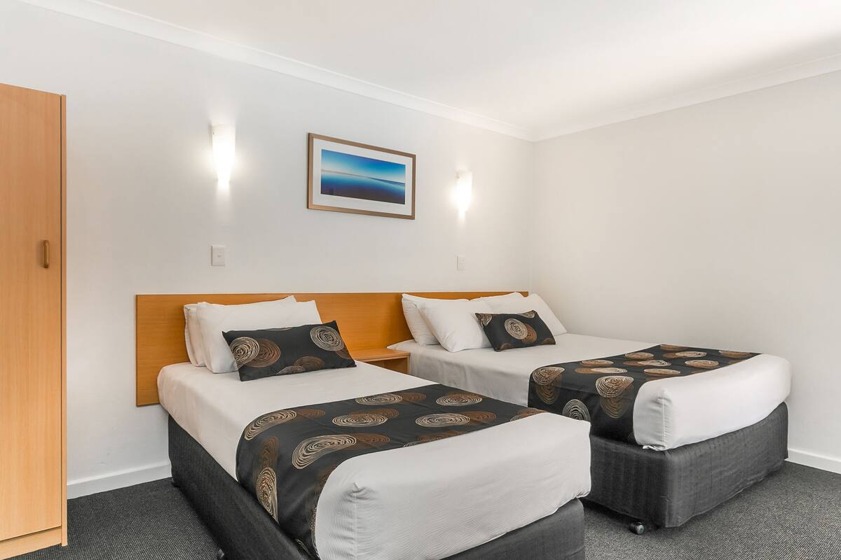 A hotel room with two single beds, a wooden headboard, a framed picture on the wall, and a carpeted floor.