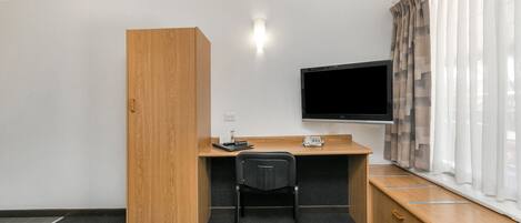 Habitación de hotel con un escritorio de madera, una silla negra, televisor, armario y ventana con cortinas.