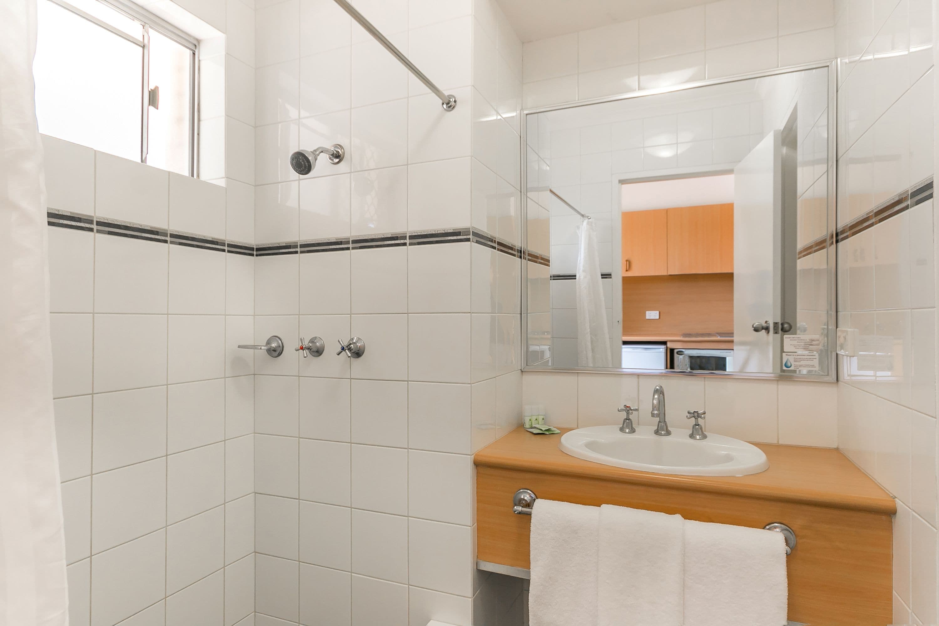 Un baño con azulejos blancos, un mueble de tocador de madera, un lavabo, un espejo y una zona de ducha.