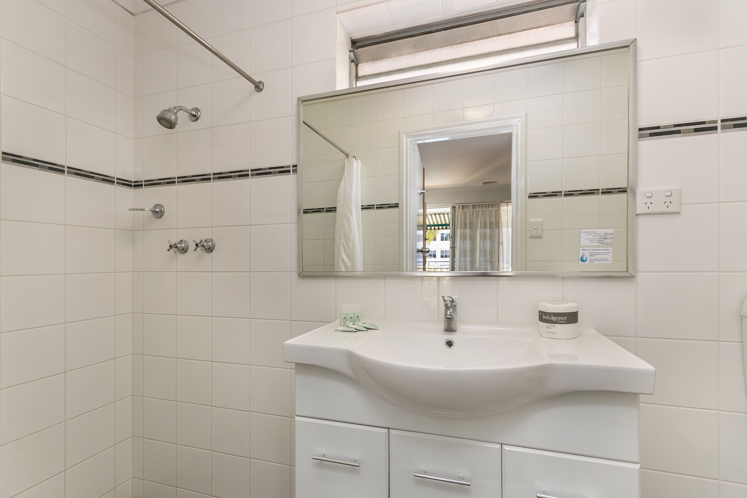 Un baño con azulejos blancos, un lavabo, un espejo, una ducha y un perchero para toallas.