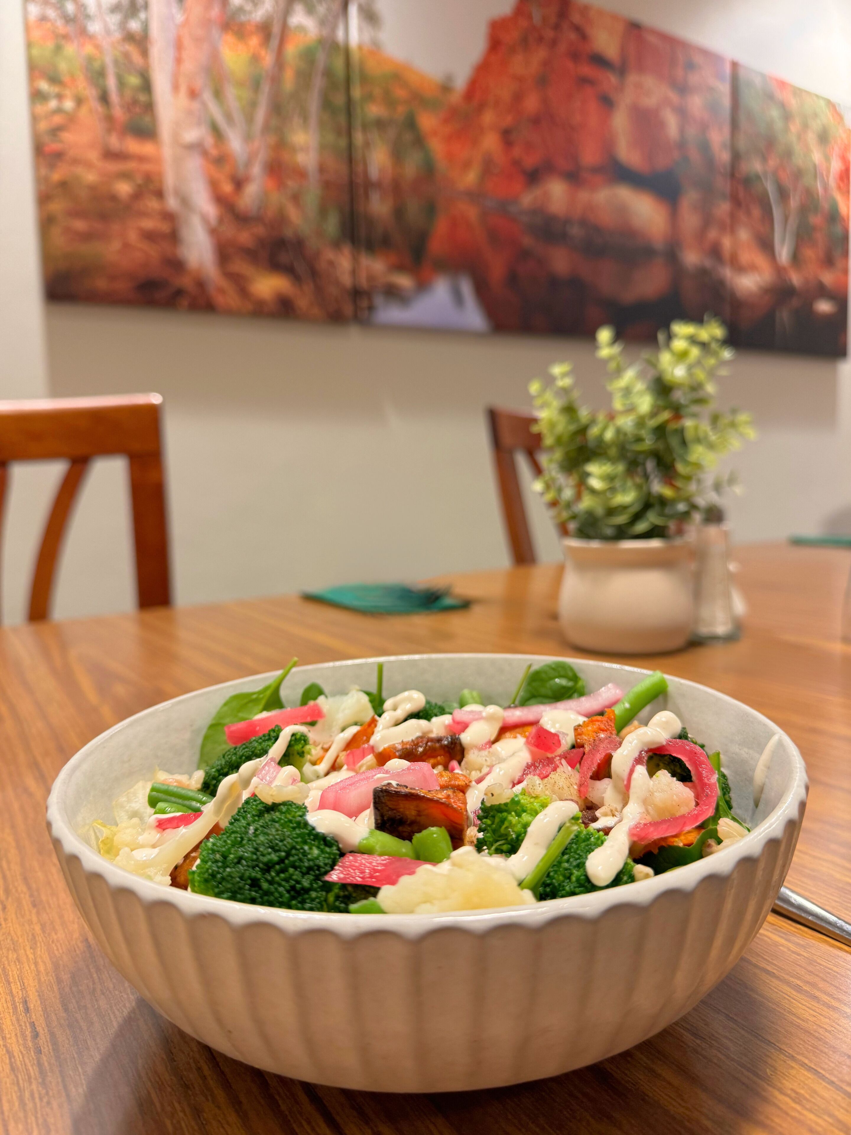 A bowl of salad on a wooden table with a painting in the background.