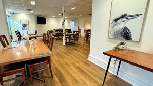 Un comedor amplio con mesas y sillas de madera, un pilar blanco con plantas y un cuadro de aves en la pared.