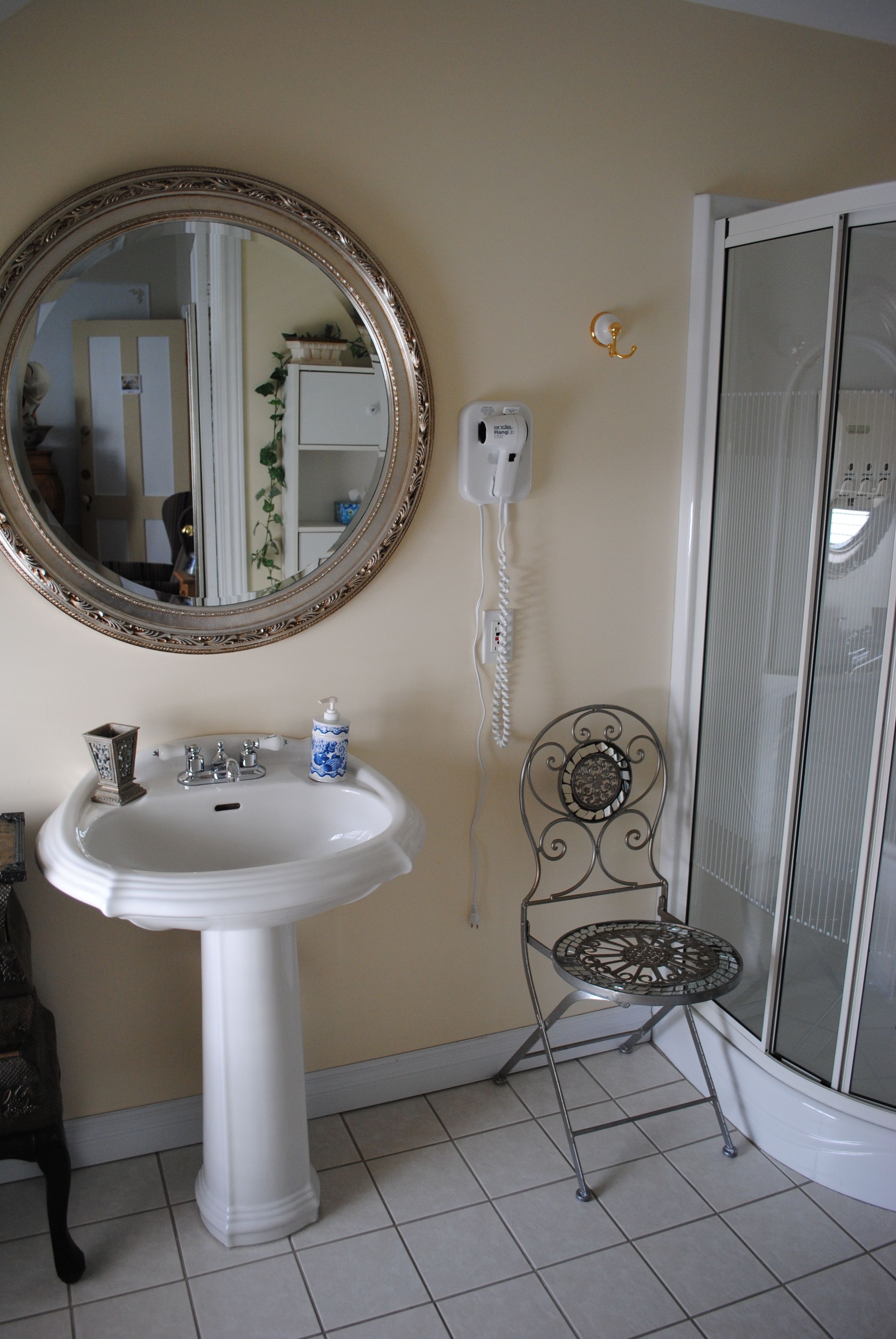 A bathroom with a pedestal sink, a round mirror with an ornate frame, a telephone, and a metal chair.