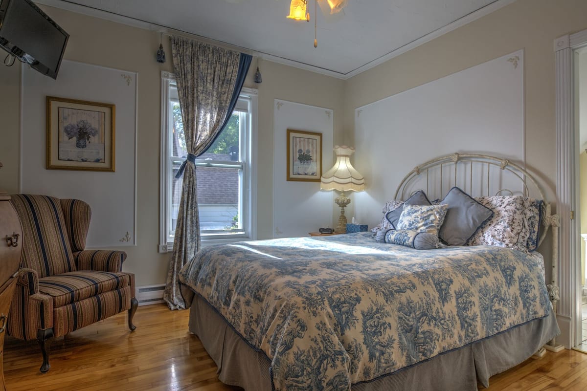 Ein Schlafzimmer mit einem großen Bett, einem Sessel, einer Lampe und zwei fensterhohen Vorhängen.