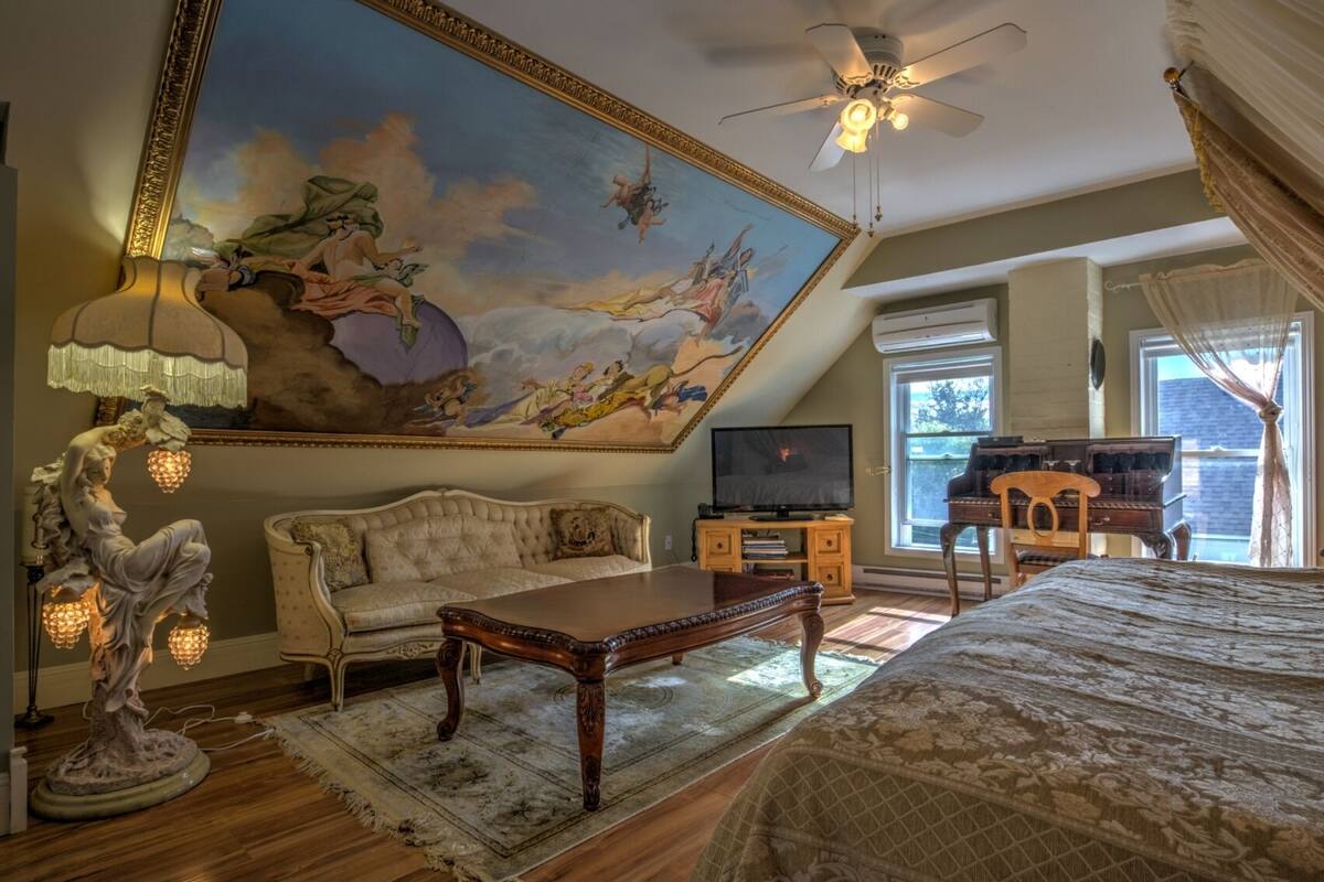 Ein geräumiges Zimmer mit einer großflächigen Deckenmalerei, einem bequemen Sofa, einem Holztisch, einem Fernseher und einem Klavier.
