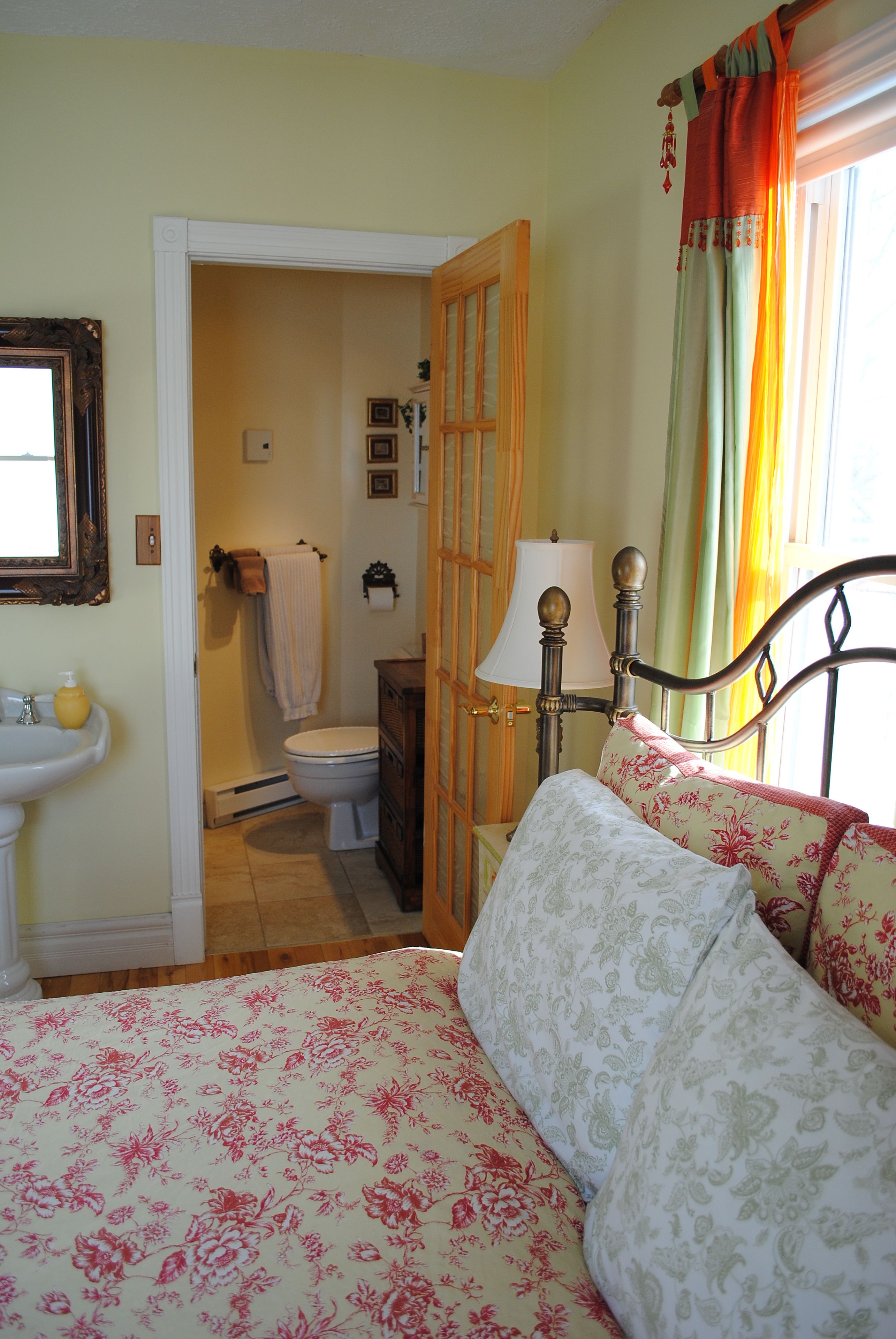 Un dormitorio con cama, mesita de noche con lámpara, ventana con cortinas, baño con inodoro y lavabo, y un espejo en la pared.