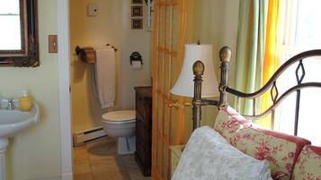 Un dormitorio con cama, mesita de noche con lámpara, ventana con cortinas, baño con inodoro y lavabo, y un espejo en la pared.