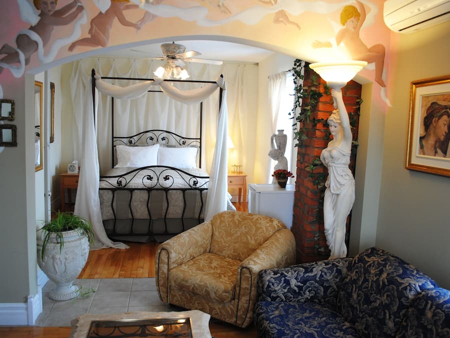 A bedroom with a four-poster bed, a sitting area with a sofa and armchair, and a decorative ceiling.