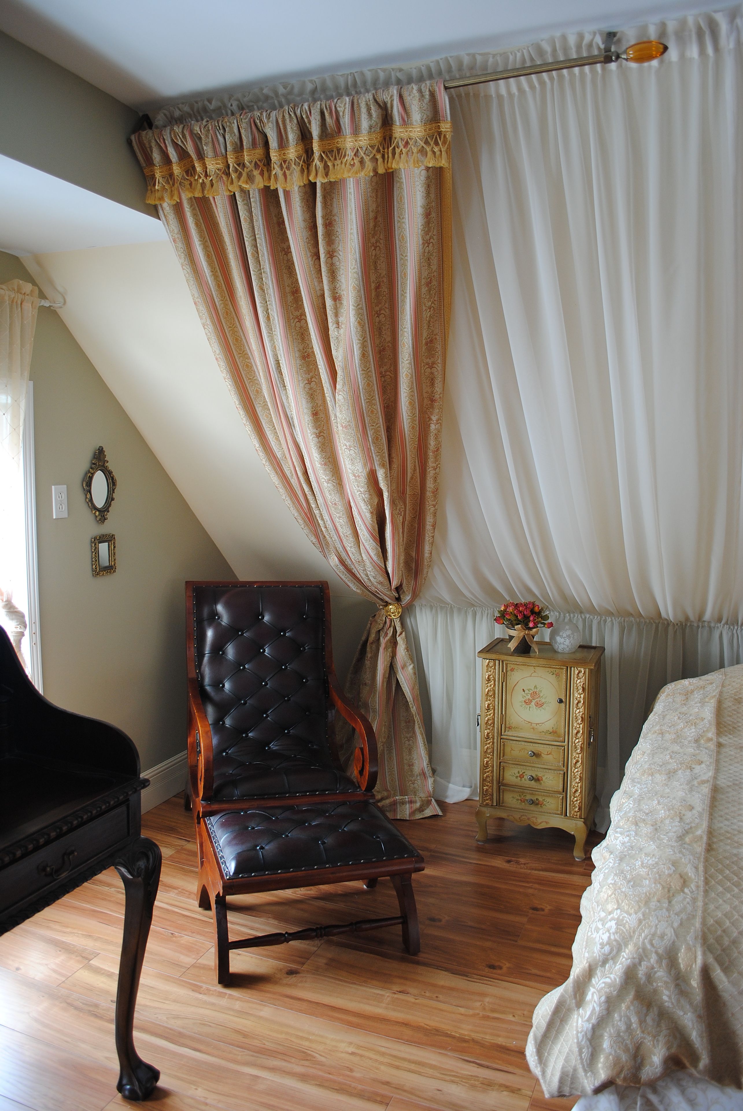 A room with a wooden floor, a black leather chair, a wooden nightstand with a vase of flowers, and a bed with a white bedspread.