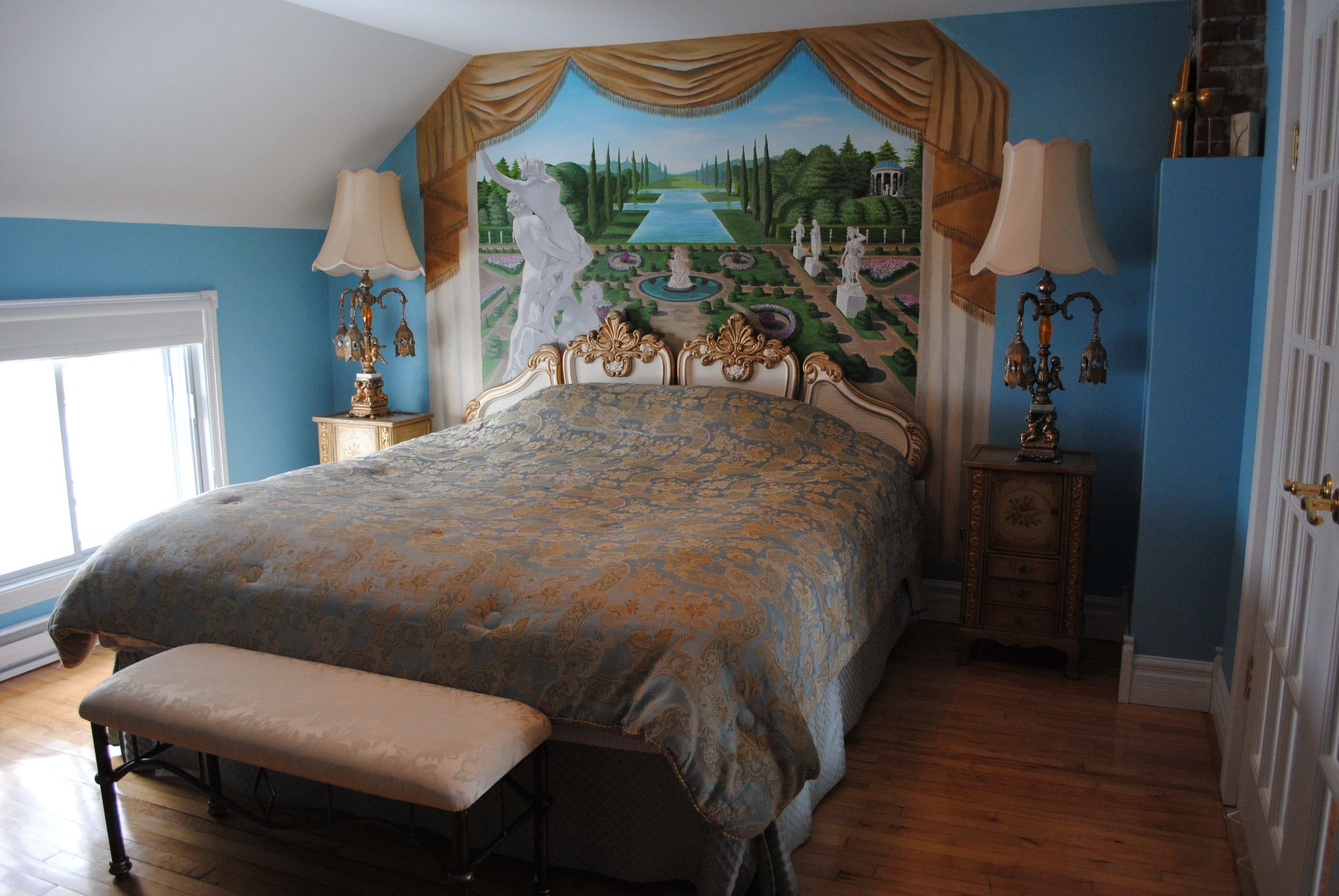 A bedroom with a large bed, a bench, a mural on the wall, and two nightstands with lamps.