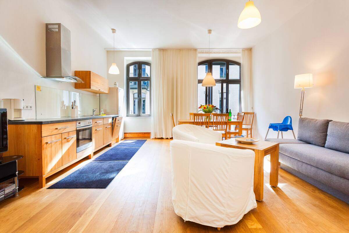 A modern kitchen with wooden cabinets, a dining area with a table and chairs, a sofa, and a large window with curtains.